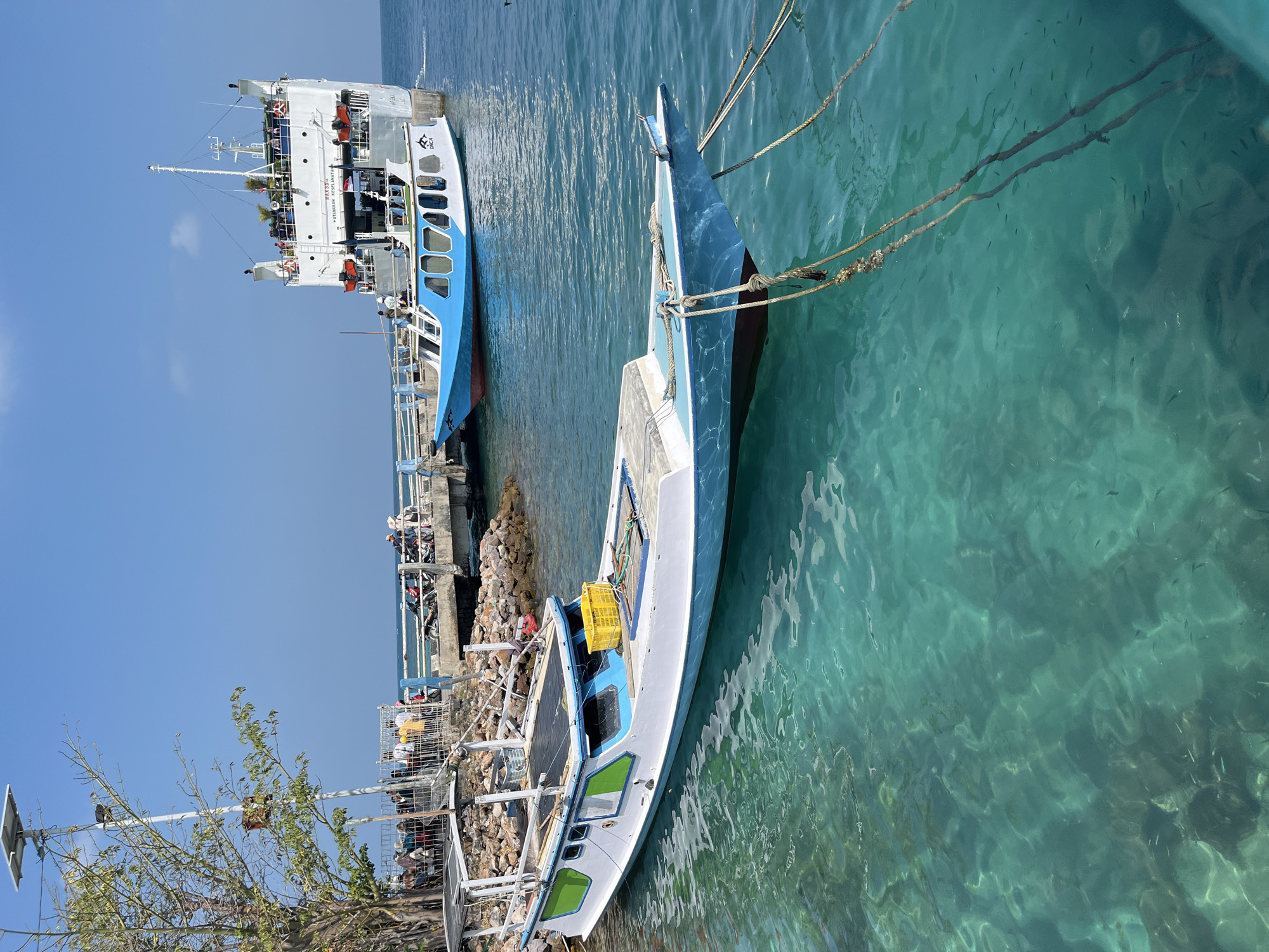 The image depicts a harbor scene in Karimunjawa, Indonesia. In the foreground, a white and blue wooden boat is moored to a pier. The boat has a covered section with a black tarp and a yellow crate on its deck. The water is clear and turquoise, revealing small fish swimming below. In the background, a larger ferry, painted white and gray, is docked at the pier. Several people are visible on the ferry and on the pier, some of whom are standing or sitting near motorcycles. A solar-powered lamp post with a camera is situated on the left side of the image, near a tree with sparse green leaves. The sky is clear and blue. The scene appears to be a busy transportation hub in the island archipelago.