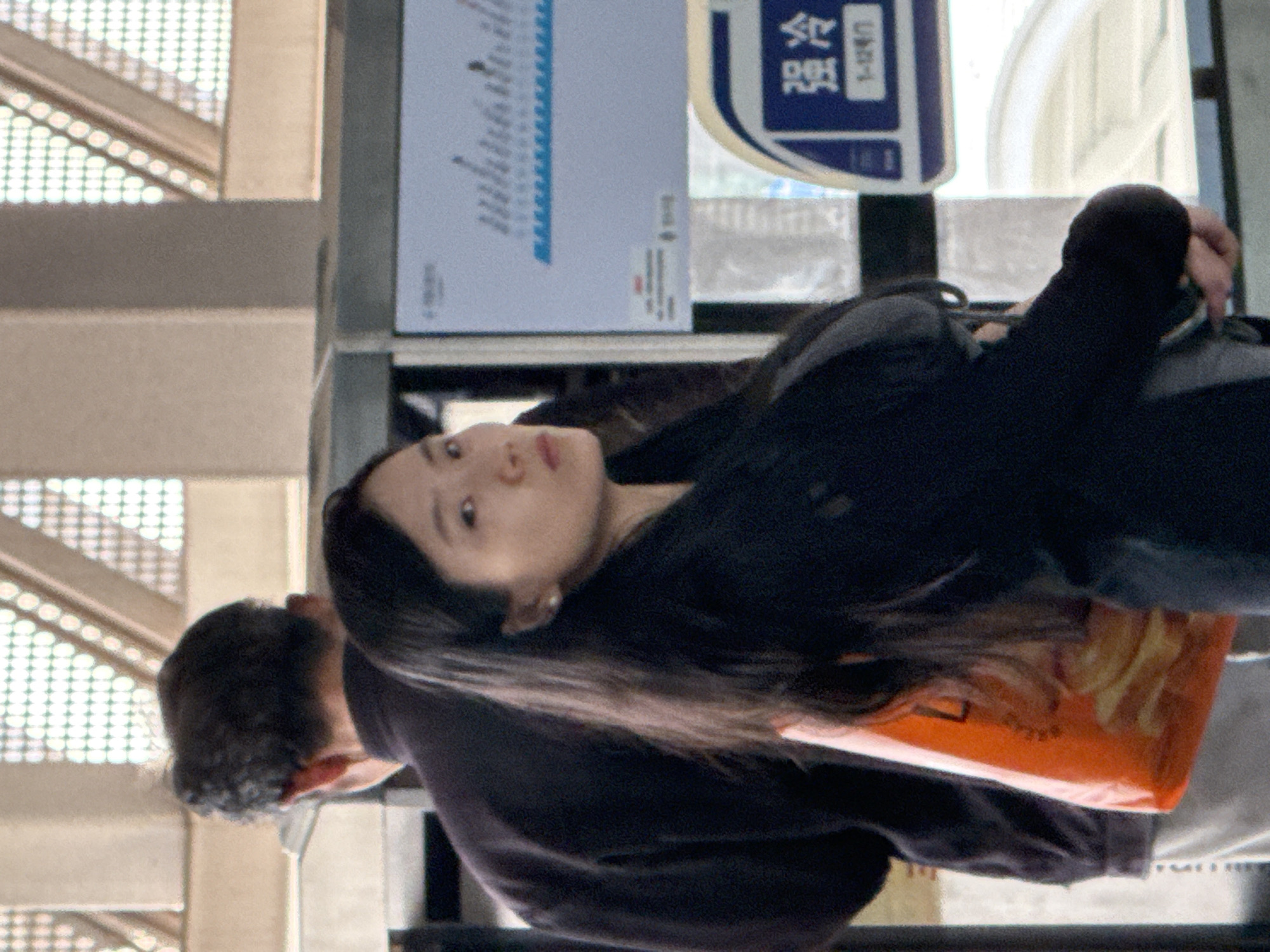 This image, rotated 90 degrees clockwise, depicts a young East Asian woman and a partially visible man in what appears to be a public indoor space in Jiangjin, China.
The woman is prominent in the foreground, looking upwards and slightly towards the viewer with a neutral expression. She has long, straight dark hair and is wearing a dark, long-sleeved top or jacket. A small, light-colored stud earring is visible on her left ear. Behind and to her right, the head and shoulder of a man are seen; he has short, dark, curly hair and also wears a dark top.
In the background, to the left of the woman, a light-colored building structure with a grid-like pattern suggests a modern interior, possibly part of an escalator or building facade. To her right, a blue and white sign prominently displays Chinese characters, including "冷" (lěng, meaning "cold"), and a partial character that could be "空" (kōng), potentially indicating "Air Conditioning" (空调). Below this, the numbers "1-1249" are visible. Another light-colored sign or display board further left shows a blurry chart or diagram with unreadable text.
An orange bag with yellow/orange patterns and some unreadable reddish-brown Chinese text is visible near the woman's lower body, suggesting it's either held by her or resting beside her. The scene is brightly lit, indicative of daytime indoors, possibly a large station, airport, or shopping mall.