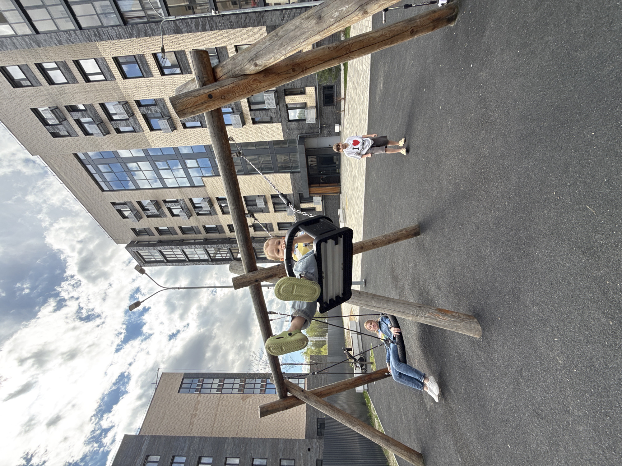The scene is a playground in Rusanovo, Russia. A wooden swing set is centrally located, occupied by two children. One child, wearing a blue denim jacket and jeans, is sitting on a tire swing. Another child is sitting on a bucket swing, wearing a yellow shirt. A third child is standing in the background, wearing a white shirt with a red heart. The playground has a dark gray surface, possibly rubber or asphalt. Two buildings are visible in the background: one with a tan brick facade and many windows, and another with a gray brick facade. A street lamp stands near one of the buildings. The sky is partly cloudy.