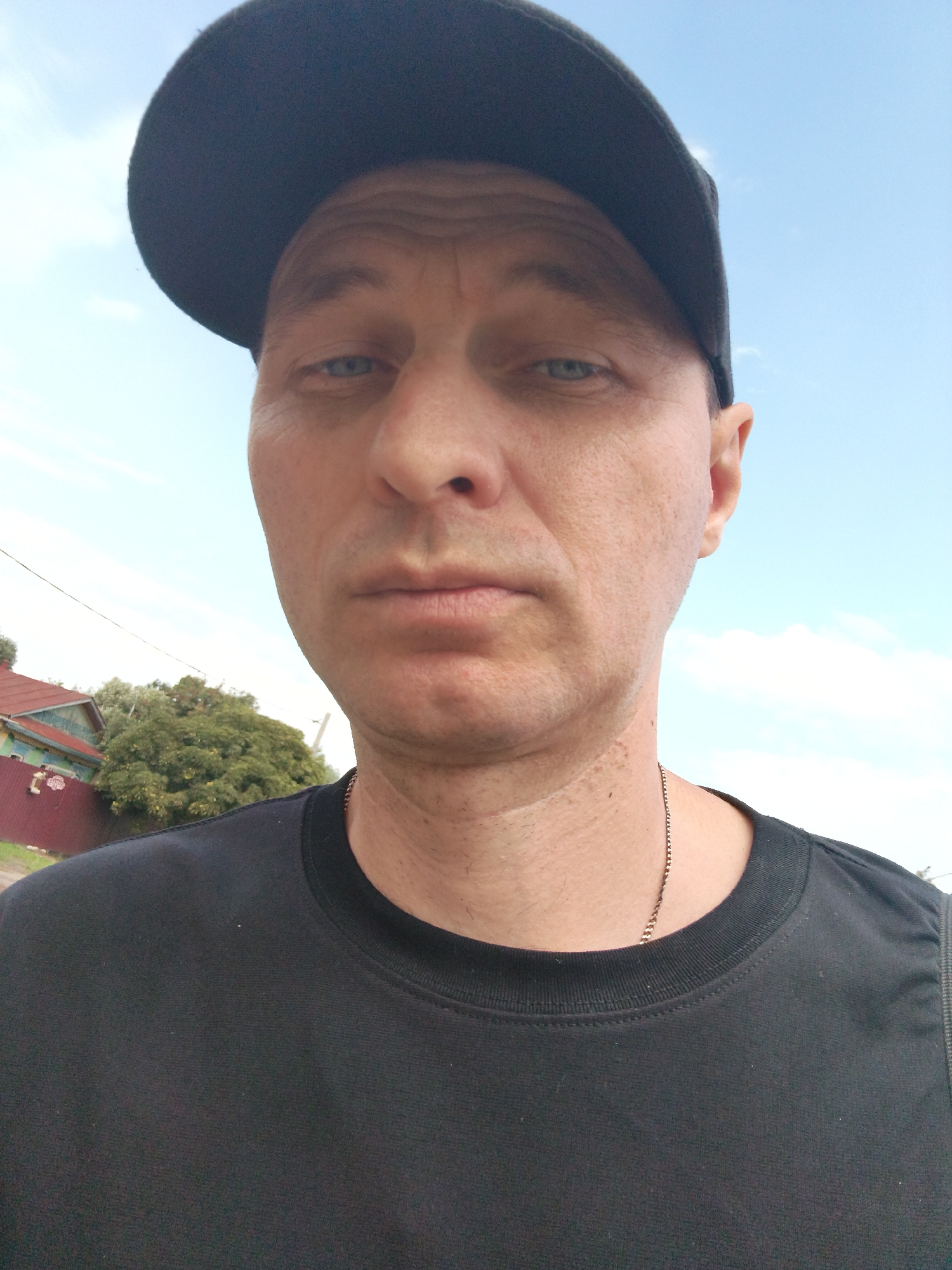 In the city of Mokshan, Russia, a man wearing a black baseball cap and a black t-shirt poses for a selfie outdoors. He has light blue eyes and fair skin. In the background, a red and green house with a fence is visible, surrounded by lush green trees. The sky is a clear blue with a few white clouds. The man's face is close to the camera, and he appears to be looking directly at it. His expression is neutral. A thin silver chain is visible around his neck.