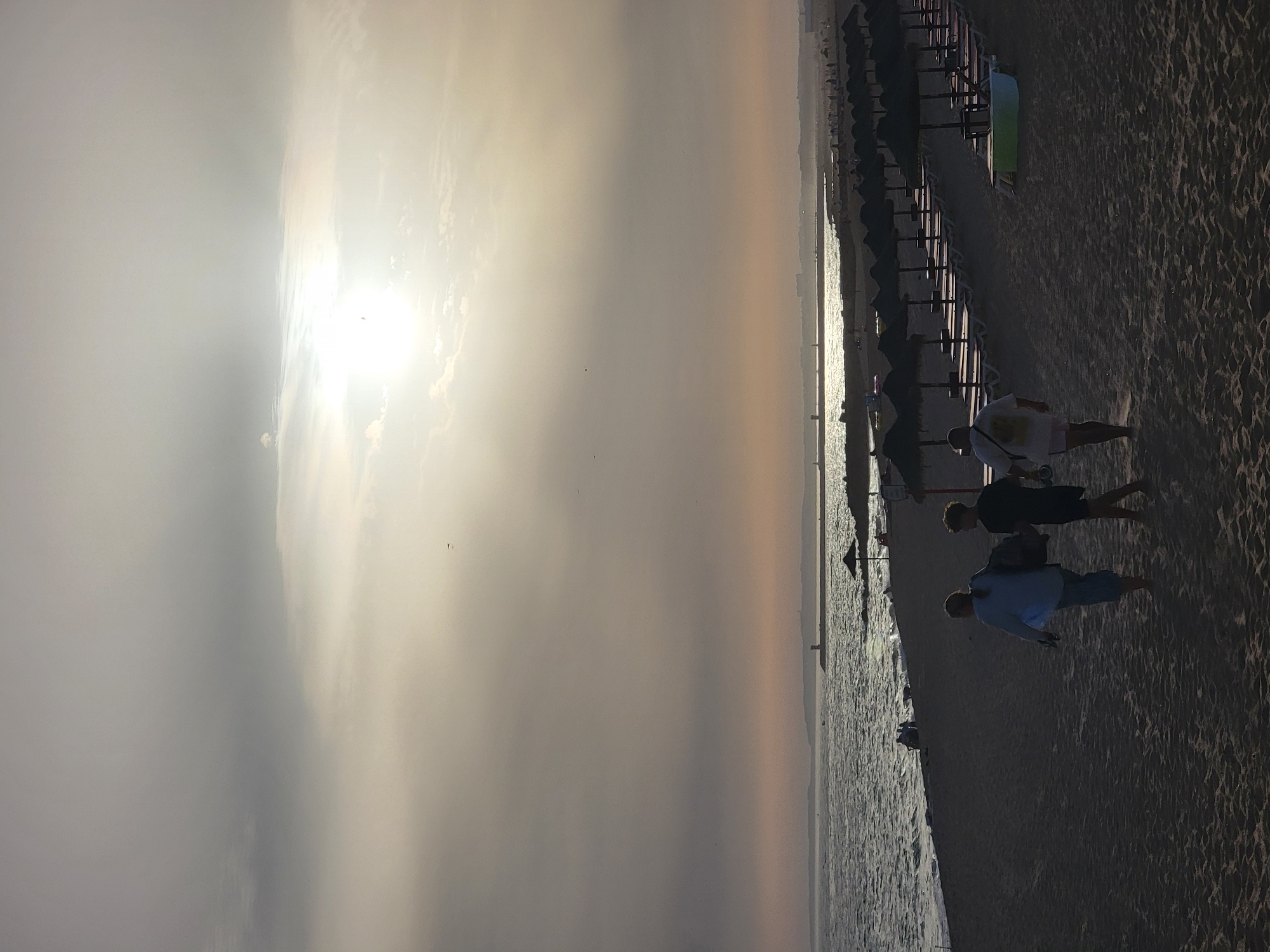 The image depicts a wide sandy beach in Quarteira, Portugal, during a hazy sunset. The sun is a bright, diffused orb in the upper right quadrant of the sky, casting a soft glow over the scene. The sky is filled with a layer of thin, wispy clouds that obscure the sun's direct light, creating a muted, atmospheric effect. In the distance, along the coastline, a lighthouse stands as a prominent landmark.
In the foreground, three figures are walking away from the camera, heading towards the water. The leftmost person, silhouetted in the dim light, appears to be wearing a light-colored top and dark pants, carrying a bag over their shoulder. To their right, a second person, also in silhouette, is wearing dark shorts and a dark t-shirt. The third figure, on the far right, is wearing a white t-shirt and light-colored shorts, with a bag slung over their shoulder. Their stances suggest a leisurely stroll along the beach.
Further down the beach, a row of thatched umbrellas and lounge chairs are neatly arranged, indicating a resort or public beach setup. Some of these chairs appear occupied by unseen individuals. The ocean stretches out to the horizon, its surface shimmering with the reflected light from the sky. Gentle waves lap against the shore, leaving a wet sheen on the sand. The overall impression is one of tranquility and the end of a day at the beach.
