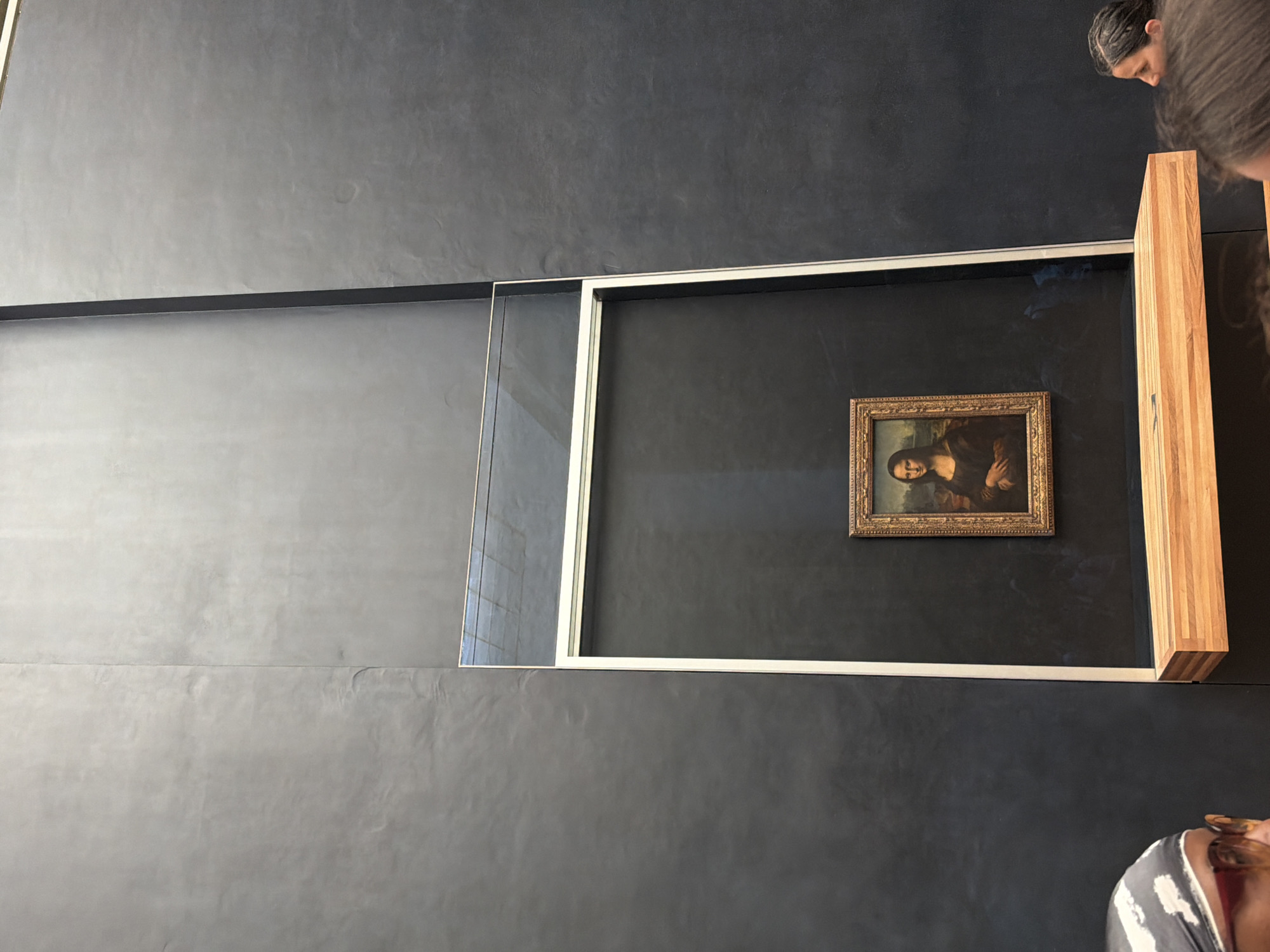 The image depicts the Mona Lisa, a 16th-century oil painting attributed to Leonardo da Vinci, housed in the Louvre Museum in Paris, France. The painting, encased in a protective glass case and framed, is displayed against a dark wall. Sections of the room with the painting are visible around it. People appear in the foreground and upper right corner, as well as in the mirror.