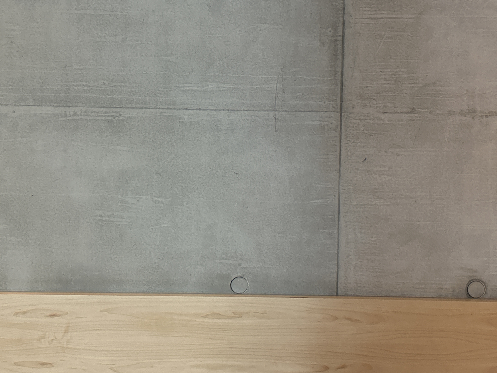 The image displays a close-up, static view of an architectural detail, presenting a clean and minimalist aesthetic. The upper two-thirds of the frame are dominated by a grey, concrete-like wall, which exhibits subtle horizontal striations and textured imperfections, suggestive of board-formed concrete or a similar finish. A distinct vertical seam or panel line is visible along the right side of the wall, running from top to bottom. A thinner, fainter horizontal line also stretches across the mid-left portion of the concrete wall.
The bottom third of the image features a smooth, light-colored wooden surface, possibly a countertop, shelf, or panel, which abuts the base of the concrete wall. This wooden surface displays natural wood grain patterns, adding warmth and contrast to the cool grey of the wall.
Resting on the wooden surface, precisely at the base of the concrete wall, are two small, circular, metallic objects. One is positioned slightly left of the center, and the other is further to the right, close to the vertical seam in the concrete. These objects appear to be small rings or caps, possibly made of a silver-toned metal.
No people or activities are depicted in the image. The lighting is even and diffused, characteristic of an indoor environment, making it impossible to determine the time of day or external weather conditions. There is no visible text. While the location context is London, United Kingdom, the image itself does not contain any specific visual cues, landmarks, or details to confirm this geographical setting. The scene focuses purely on the textures and forms of the building materials.