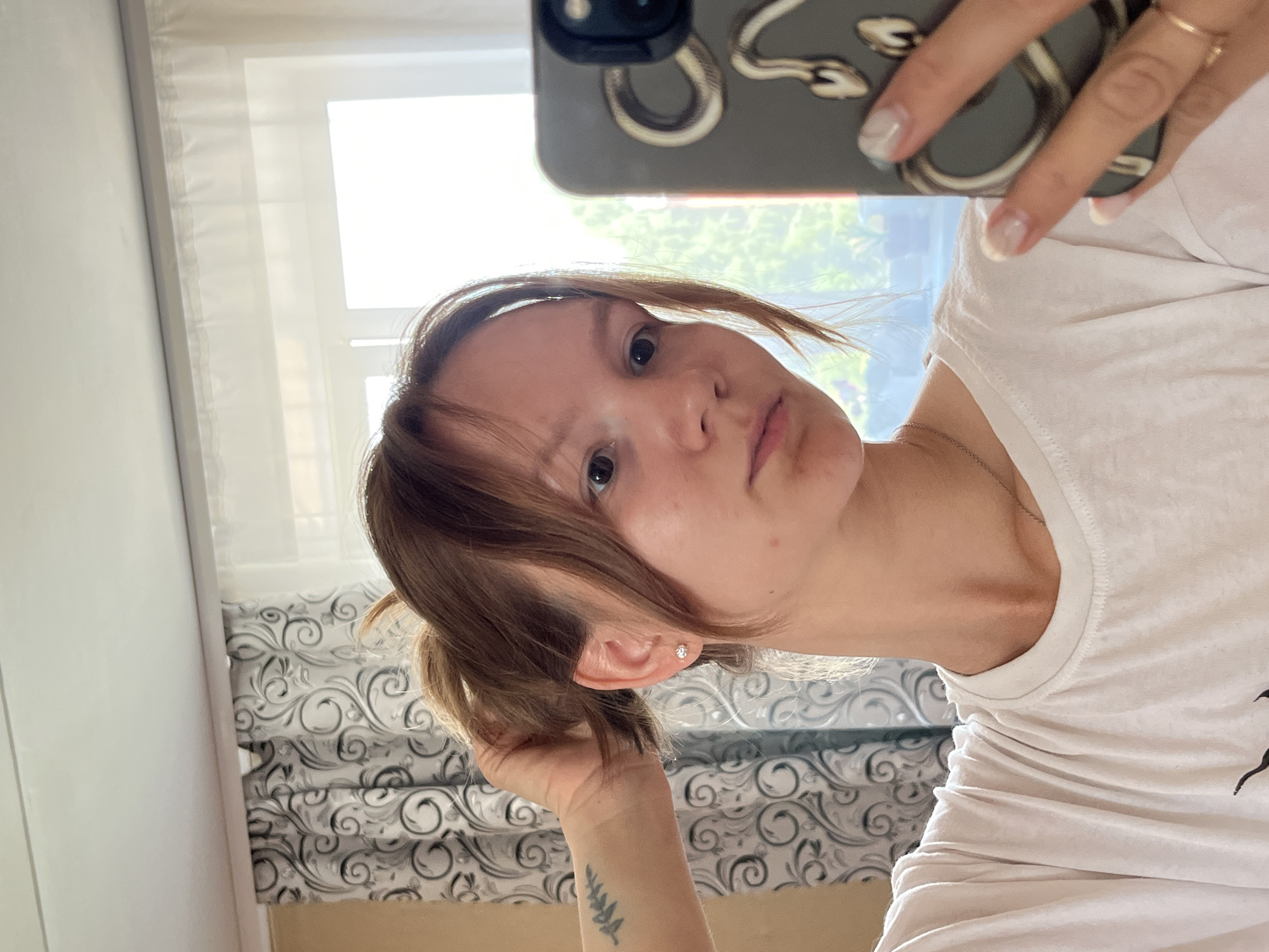 A young Caucasian female is captured in a mirror selfie within what appears to be a residential setting, possibly a bedroom, in Noviy, Russia. The woman has light brown hair pulled back into a messy ponytail with loose strands framing her face. She is wearing a plain white t-shirt and a delicate silver necklace. Her right hand is raised, adjusting her hair, while her left hand holds a smartphone with a dark grey case featuring a snake design. The background shows a window with sheer white curtains and patterned grey curtains. The natural light suggests it is daytime. The overall scene is casual and personal.