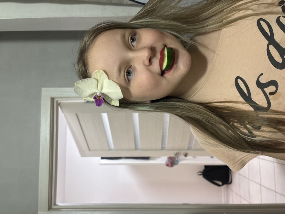 The image, rotated 90 degrees counter-clockwise, features a young girl (appearing to be a pre-teen or early teen) posing playfully in an indoor setting. She has light-colored eyes and long, straight hair that is light brown or ash blonde, with subtle hints of a greenish or bluish tint in some strands. A white orchid with a distinctive purple center is tucked into her hair above her left ear, resting on her forehead. She is wearing red lipstick, and a slice of green cucumber is held between her lips, creating a humorous, wide-mouthed, and quirky expression. Her overall demeanor appears cheerful and mischievous.
She is wearing a light peach or tan colored t-shirt. On the front of the t-shirt, there is black cursive text. While most of the text is obscured by her hair or is out of frame, the word "is" is clearly visible, preceded by the beginning of another word that appears to start with "L".
The background shows an indoor environment. To the left of the girl's head, a plain grey wall is visible. To her right and behind her, there is a light-colored wooden door with multiple vertical slats or panels, which is partially open. Through the opening of the door, a white wall is visible further back. In the distance, a black backpack is resting on what appears to be the floor, and some other indistinct items are visible on a surface or shelf. The lighting is consistent with an indoor setting, likely artificial. The photograph captures a fun and lighthearted moment.