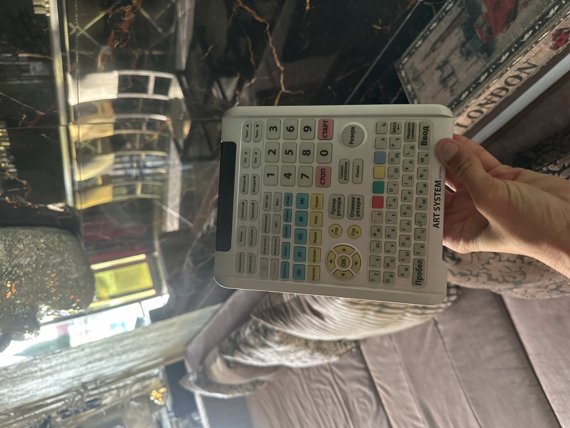 A person is holding a white remote control in their right hand, with their fingers positioned around the bottom half of the device. The remote has a keypad with numbers and letters, along with various function buttons. The background is blurred, but appears to be an indoor setting with furniture that suggests a living room or lounge area. There are elements of decor visible, including what looks like a mirrored surface with a marble-like pattern. The overall lighting is dim, with some light sources contributing to reflections on the surfaces.
There is no information or visual evidence in the image to determine the location within the city of Brca, Montenegro.