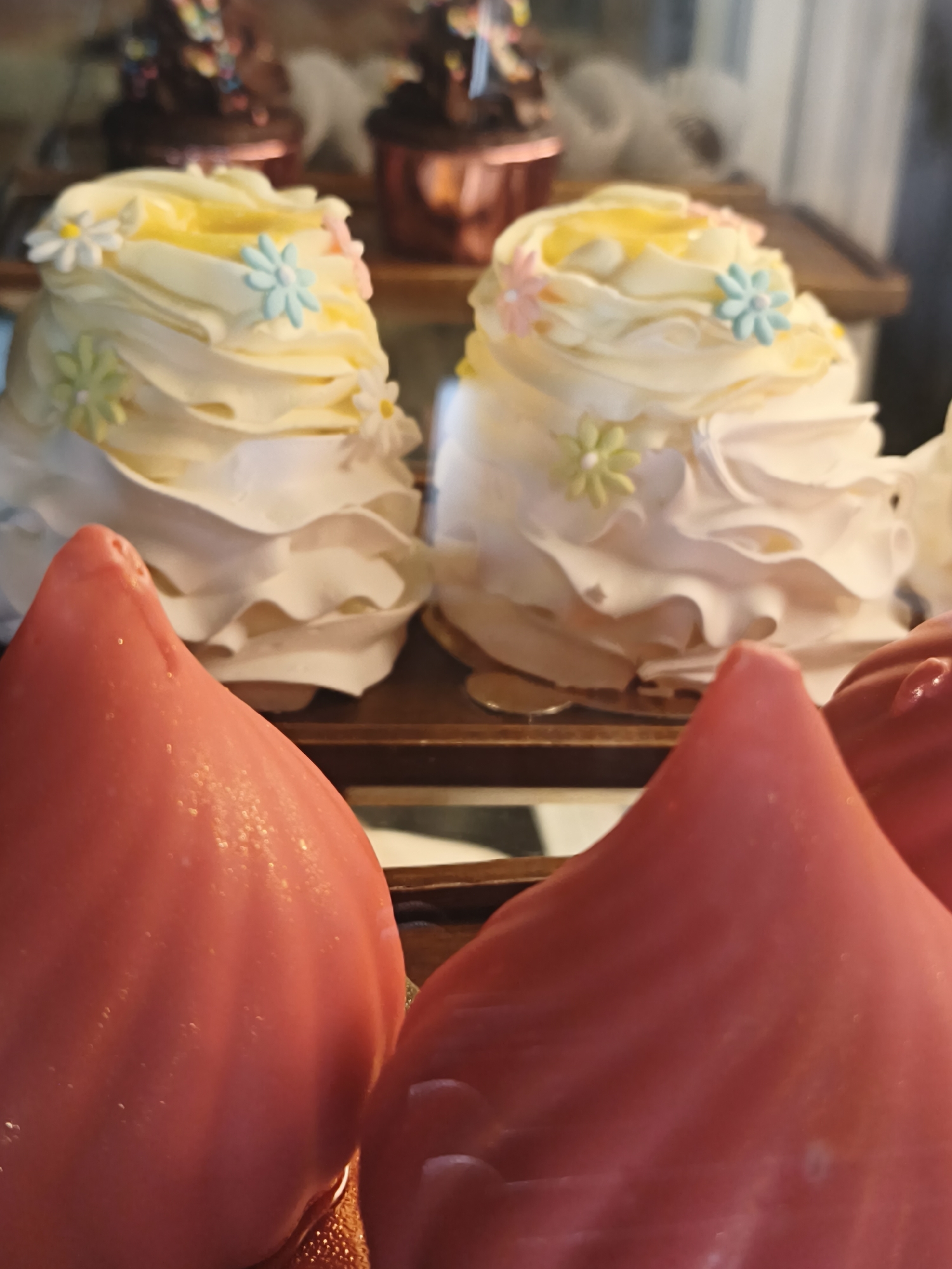 In the city of Nha Trang, Vietnam, within Ward Bac, a close-up shot focuses on two vibrant, cone-shaped desserts in the foreground, their surfaces glistening with what appears to be condensation or a glossy glaze. Behind them, two elaborately decorated cupcakes are visible. These cupcakes feature swirled white frosting with yellow centers and are adorned with small, pastel-colored edible flowers. The cupcakes are presented on a tiered display, with other desserts, some appearing to be chocolate cupcakes with colorful sprinkles, also present on upper shelves. The background is blurred, suggesting the image was taken inside a shop or bakery, possibly a display case.