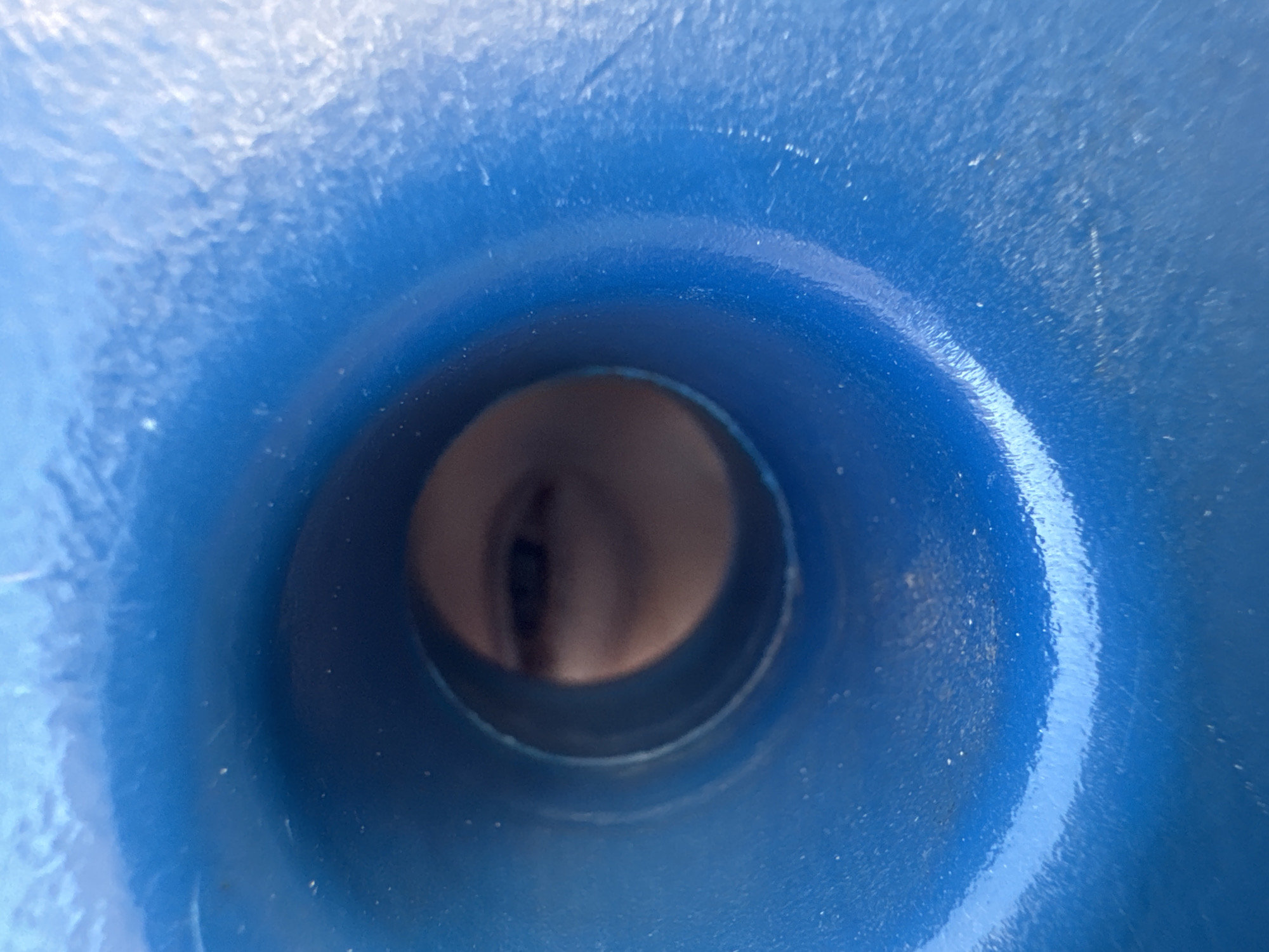 An extreme close-up shot shows an eye peering through a circular opening in a blue plastic object. The opening is roughly in the center of the frame and appears to be a tunnel or hole. The eye is partially visible and seems to be looking directly at the camera. The blue object has a slightly textured surface, and there are some specks of white or light-colored debris scattered across it. The lighting suggests the scene is outdoors, possibly on a sunny day, as there are highlights on the blue surface. The specific location in Saint-Claude, France, is not discernible from the image itself.