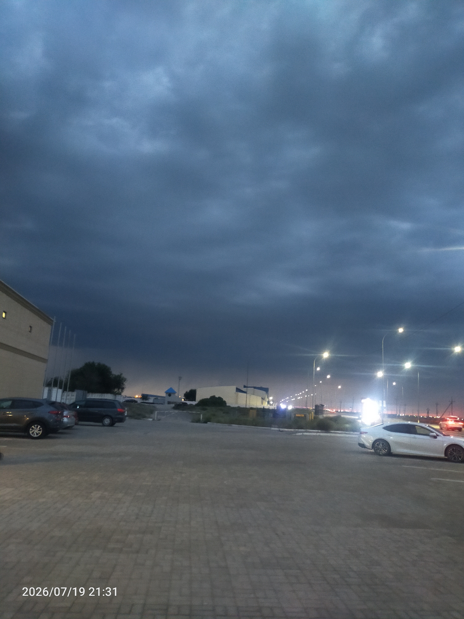 The image depicts a dark, cloudy sky over an outdoor parking lot. Several cars are visible, including a white sedan parked in the foreground and a dark SUV and a dark sedan parked to the left. A light-colored building with a yellow accent is on the left edge of the frame. Streetlights are illuminated in the distance, suggesting it is evening or night. The ground is paved with interlocking stones. The timestamp "2026/07/19 21:31" is visible in the bottom left corner of the image.
