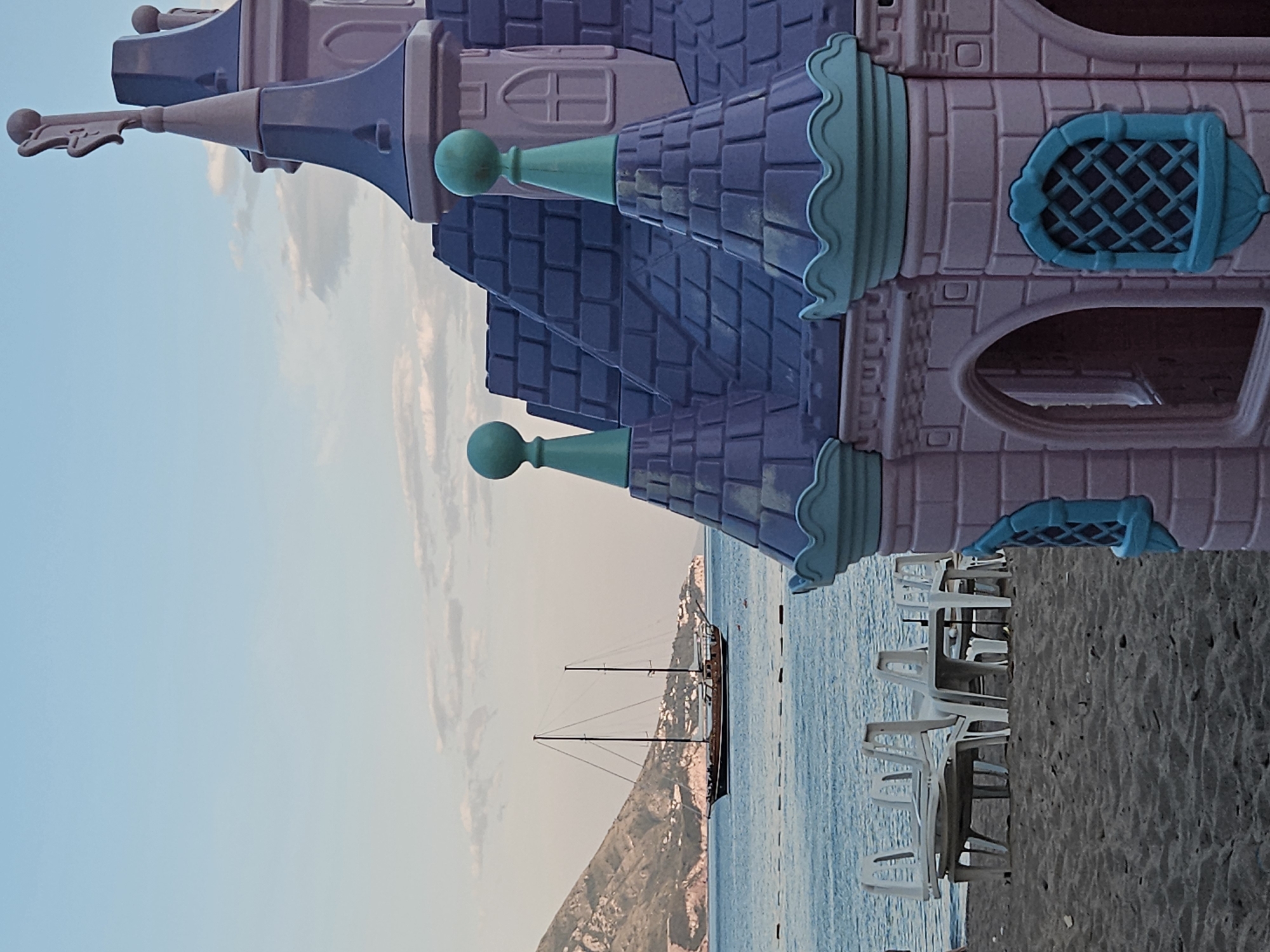 The image shows a scene from a beach, with a toy castle structure prominently in the foreground. The castle is purple and pink, with blue-green decorative elements. In the background, the scene opens to a view of the sea, where a sailboat is visible against the backdrop of a mountainous coastline. The beach itself is populated with rows of white chairs, likely part of a beach resort setup. The sky is a pale blue, suggesting either dawn or dusk. The image appears to be taken at an outdoor location, the beach, potentially in the city of Unknown, Unknown.