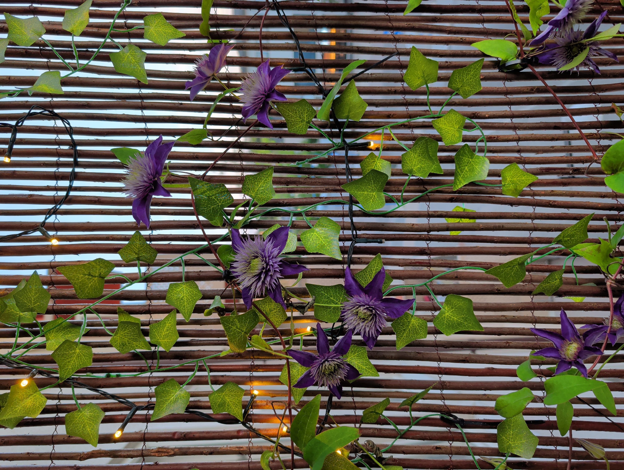 The image shows a close-up of a decorative screen made of vertical bamboo or wooden poles. Green artificial ivy leaves and purple artificial clematis flowers are attached to the screen, intertwined with strings of small, warm-toned LED lights. The background appears to be a sky with some buildings or structures visible through the gaps in the poles. The overall impression is of an outdoor decorative element, possibly on a balcony or patio. The location is Nynäshamn, Sweden. There are no people or specific actions depicted.