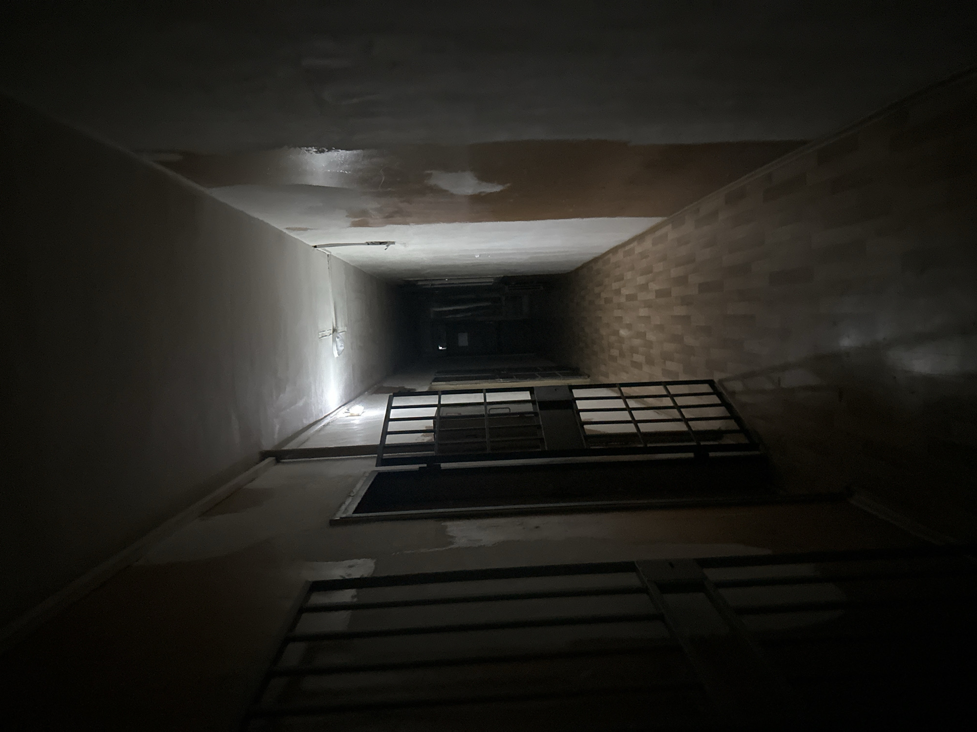 The image depicts a dimly lit stairwell, viewed from a low angle looking upwards. The walls are bare, with one side appearing to be a painted surface and the other covered in brick. The ceiling shows signs of water damage. Metal railings and grates are visible on each level, leading up into the darkness. The setting appears to be an urban environment with signs of neglect. It is not possible to definitively identify the location as Sofia, Bulgaria, from the image alone.