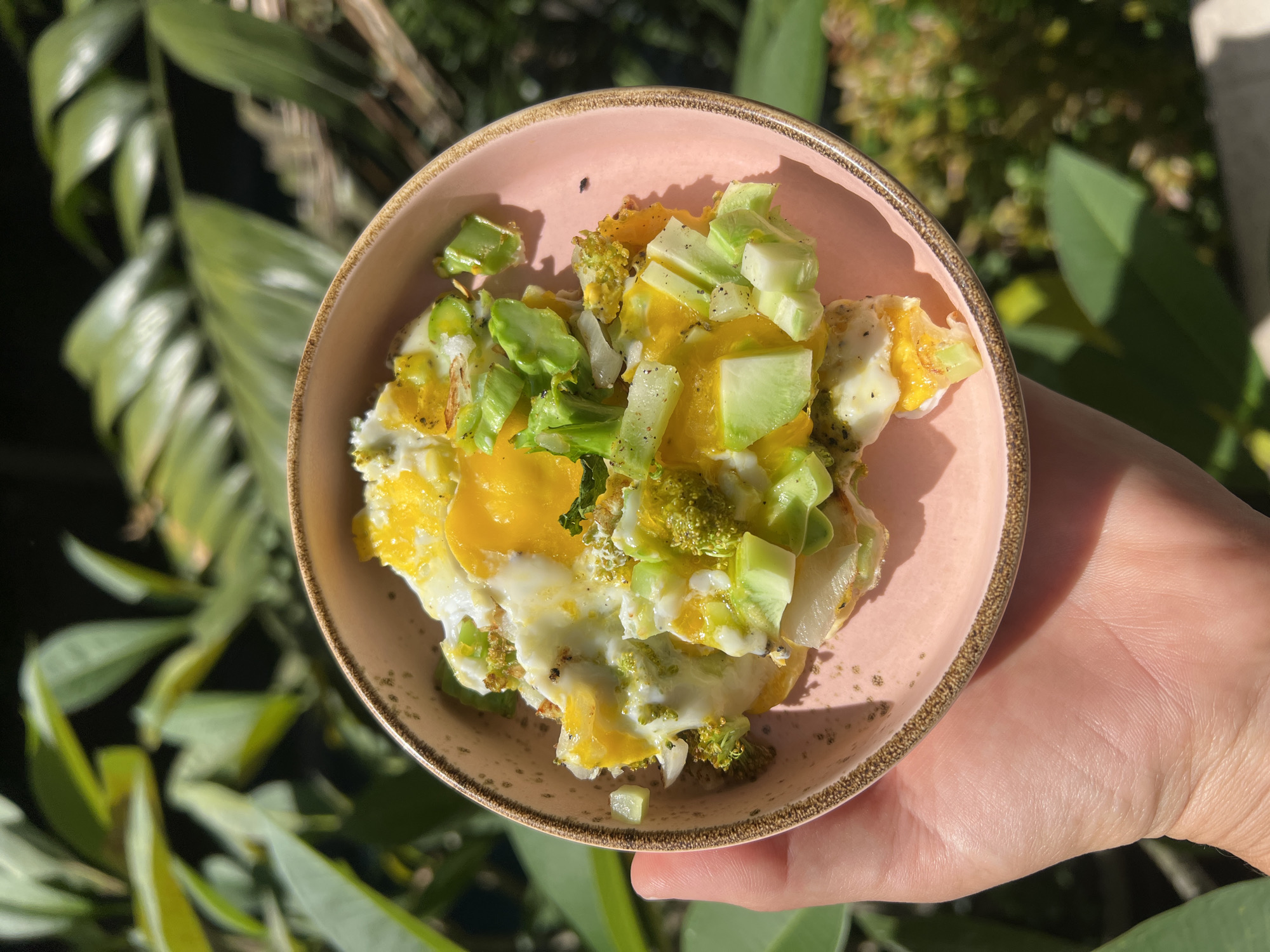 A hand is holding a pink ceramic bowl filled with an egg-based dish. The dish contains visible ingredients like diced green vegetables, possibly zucchini or a similar squash, and what appears to be broccoli florets. Parts of the egg yolk are visible, surrounded by cooked egg whites. The bowl's rim is accented with a gold-colored band. The backdrop includes dense foliage, indicative of a tropical or subtropical environment. Given the specified location of Ubud District, Indonesia, the environment is consistent with the region's lush landscapes. The overall scene is sunlit.