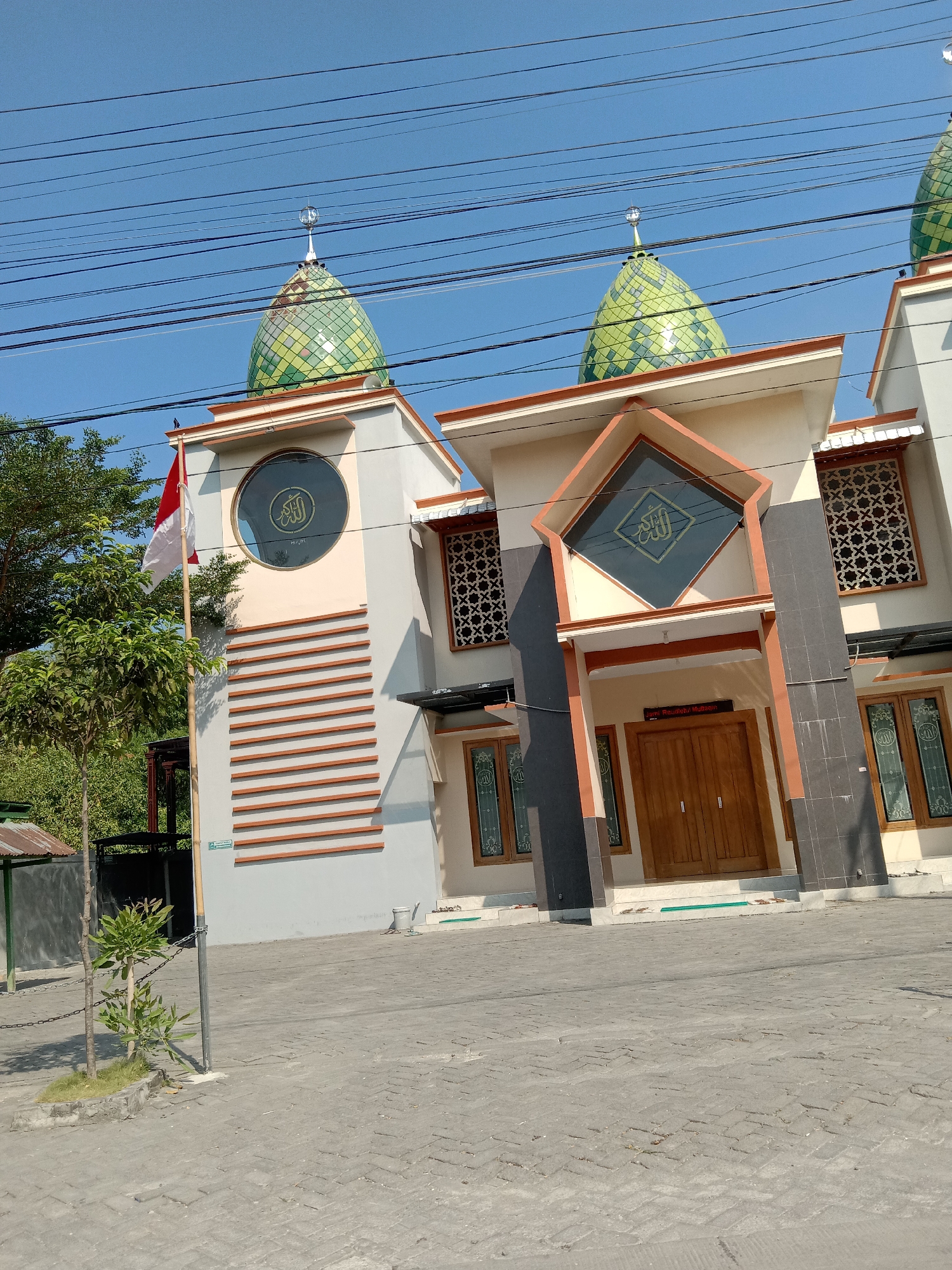 A photograph captures the exterior of a mosque in Siwalan, Indonesia, under a clear blue sky. The mosque features two prominent green, tiled domes topped with crescent moons. The building's facade is a mix of light gray, beige, and dark gray panels, with decorative patterns on some windows. An Indonesian flag flies on a pole to the left of the entrance. In front of the mosque is a paved courtyard. A small tree with green foliage is situated in the foreground. Power lines are visible crisscrossing the sky above the mosque.