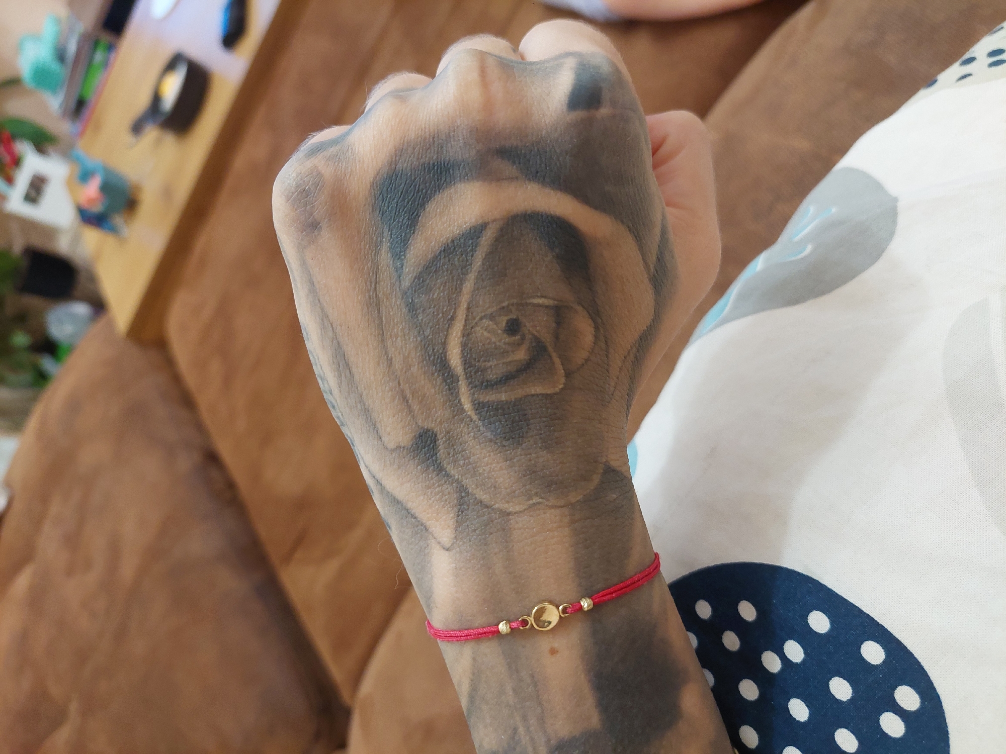 A close-up shot displays a tattooed hand, predominantly featuring a rose design on the top. The hand is adorned with a red string bracelet that has small golden beads and a circular centerpiece. The hand rests against a surface with a pattern of large blue circles on a white background. The background includes blurred furniture, potentially a couch or table, in shades of brown and hints of green. No location in Plovdiv, Bulgaria can be determined from the image.