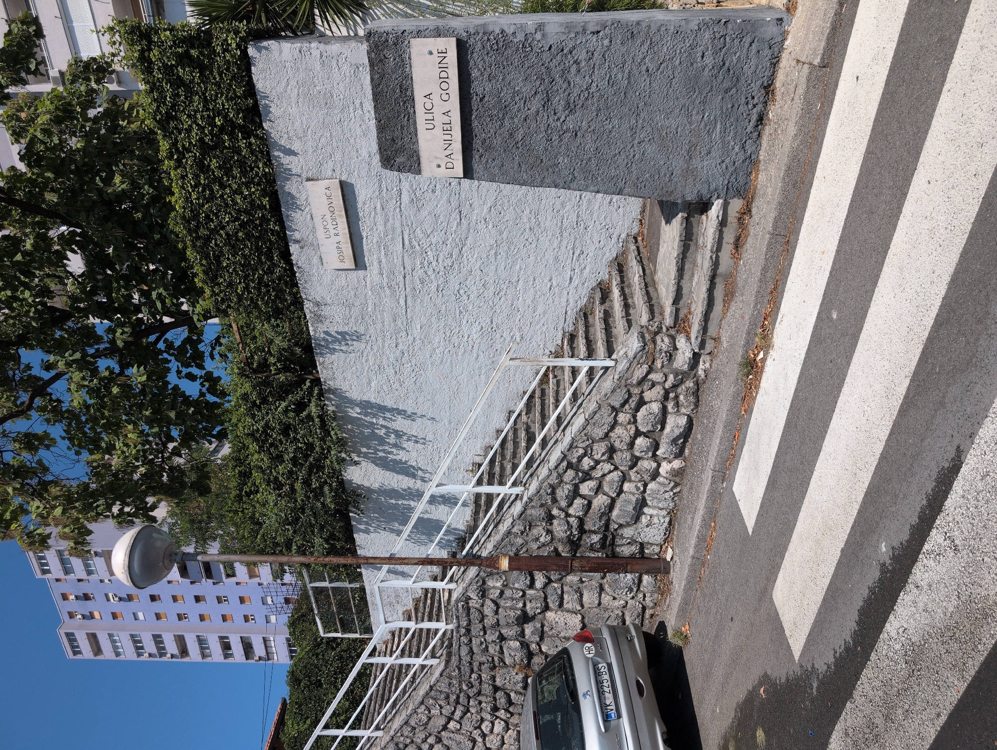 The image depicts an outdoor scene in Rijeka, Croatia, featuring a street sign indicating "ULICA DANIJELA GODINE" and another sign that reads "USPON JOSIPA RADINOVIĆA". A pedestrian crossing is visible in the foreground. To the left, a silver car is parked next to a stone-walled embankment leading to a set of stairs with white railings. A lamppost stands beside the stairs. In the background, a tall, light-purple residential building is prominent, surrounded by lush green foliage. The sky is clear and blue.