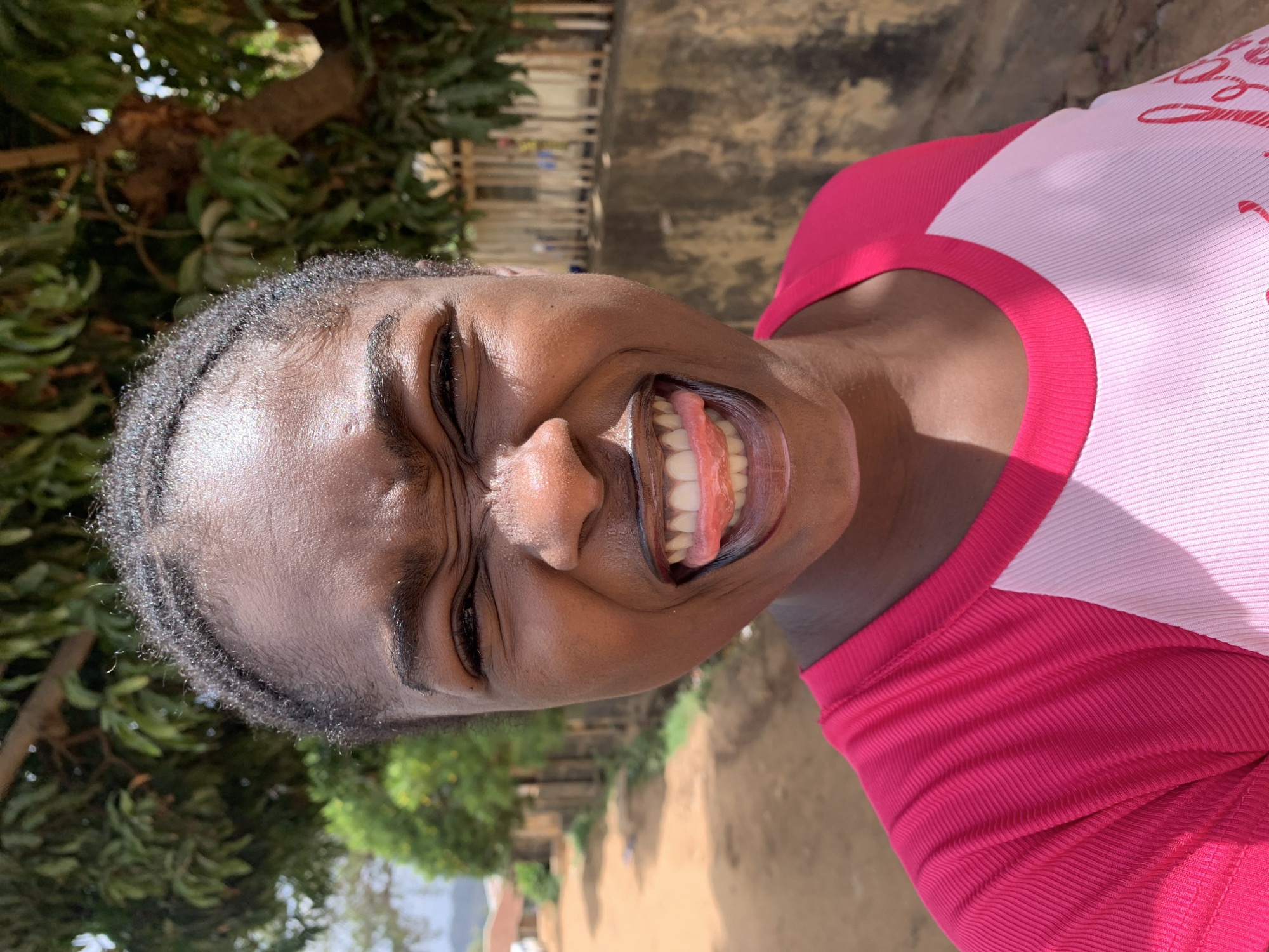 A close-up, rotated portrait shows a dark-skinned woman in Rafin Par, Nigeria, grinning widely with her eyes squinted nearly shut. She wears a pink and white t-shirt and is positioned outdoors with trees visible to the left, and a dirt path in the background leading to distant buildings, suggesting a rural or semi-rural location within the city. A fence is slightly visible behind the trees.