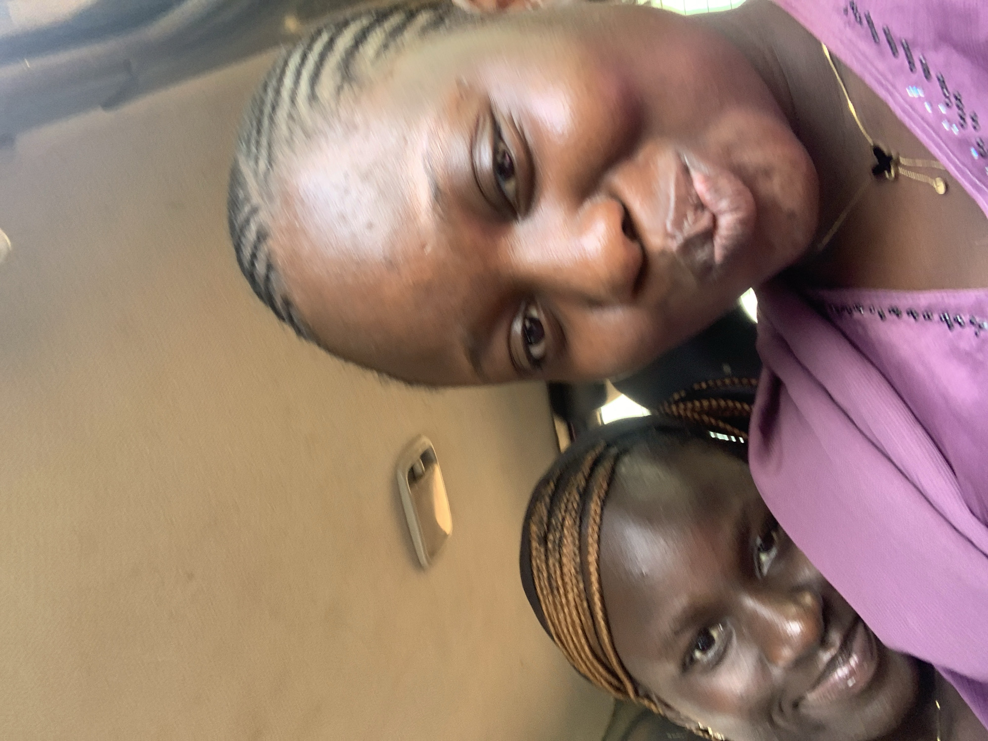 Two young Black women are inside a vehicle. The woman in the foreground is taking a selfie and is looking at the camera with her lips pursed. She is wearing a purple garment and a gold necklace with a black pendant. The woman in the background is partially visible, looking towards the camera with a slight smile. She has braids and is wearing a gold headband. The interior of the vehicle is visible, with the ceiling of the car and a light fixture. The lighting suggests it is daytime.