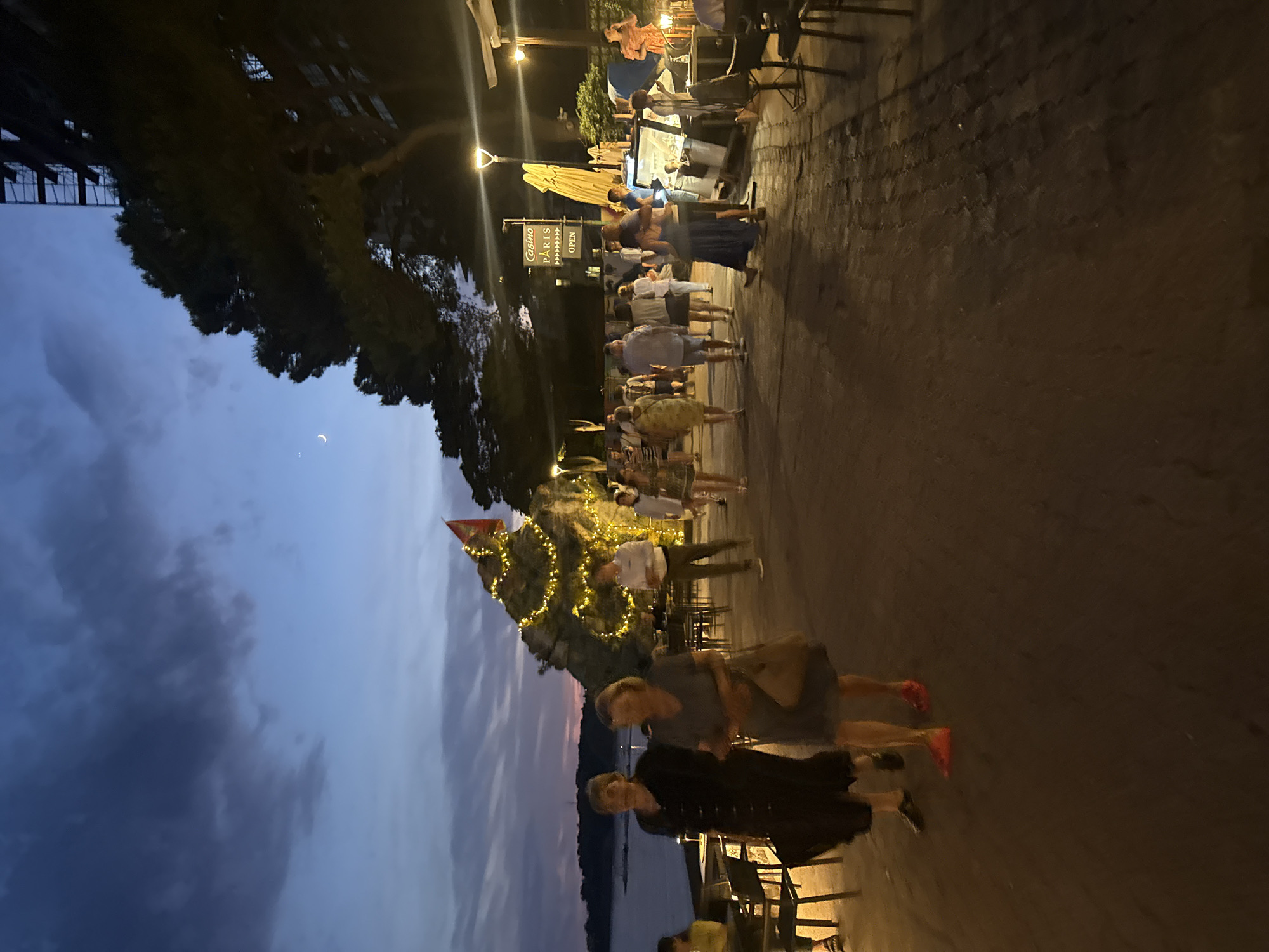 This image shows a twilight scene in Rafailovići, Montenegro, a coastal town known for its beaches and promenade. The sky is a deep blue with streaks of pink and purple, indicating the sun has recently set. A crescent moon is visible in the upper left portion of the sky. The promenade is paved with cobblestones and lined with outdoor seating areas for cafes and restaurants. People are walking along the promenade, some in groups and others individually.
In the foreground, two women are walking towards the camera. The woman on the left is wearing a dark dress, and the woman on the right is wearing a lighter colored dress. In the mid-ground, there are more people walking and standing around. To the right of the promenade, there are several outdoor dining areas with tables and chairs. In the background, a rocky outcrop is illuminated with fairy lights, and a red flag flies from the top. Further in the background, there is a building under construction with scaffolding. A sign indicating "Casino Paris Open" is visible on a pole. The sea is visible on the left side of the image, with a dark silhouette of trees and buildings along the coast. The overall atmosphere is relaxed and leisurely, typical of a summer evening in a tourist resort.