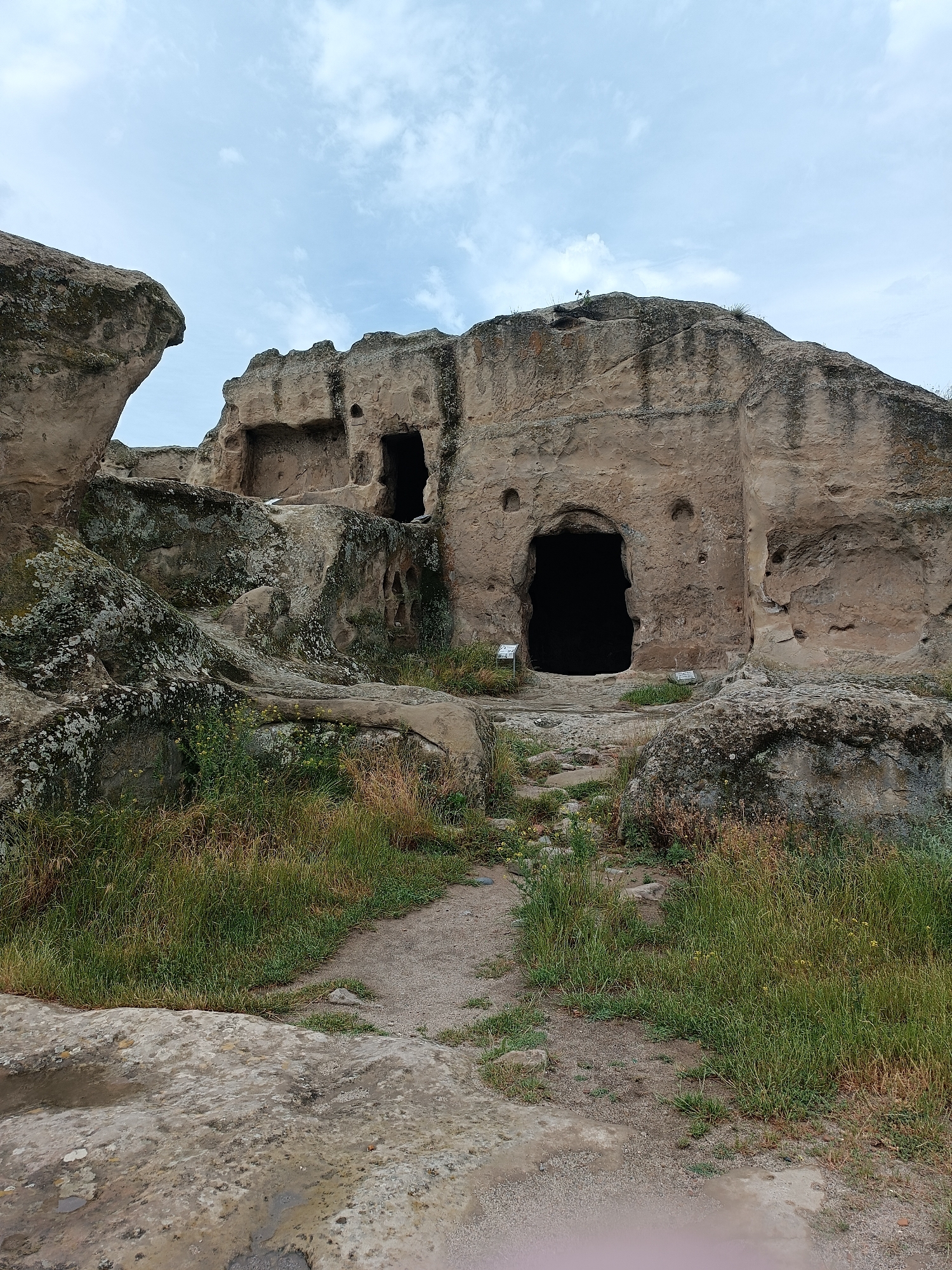 The image depicts the Uplistsikhe cave complex, a historically significant archaeological site near Gori in Georgia. In the foreground, a path made of dirt and stones leads towards the cave entrances carved into a large rock formation. Lush green grass and some dry vegetation grow on either side of the path and around the base of the rocks. The rock face is rugged and weathered, with several openings of varying sizes, some appearing as dark voids. The sky above is overcast with scattered clouds, suggesting a temperate climate. The overall scene conveys an ancient, untouched landscape with a sense of history.