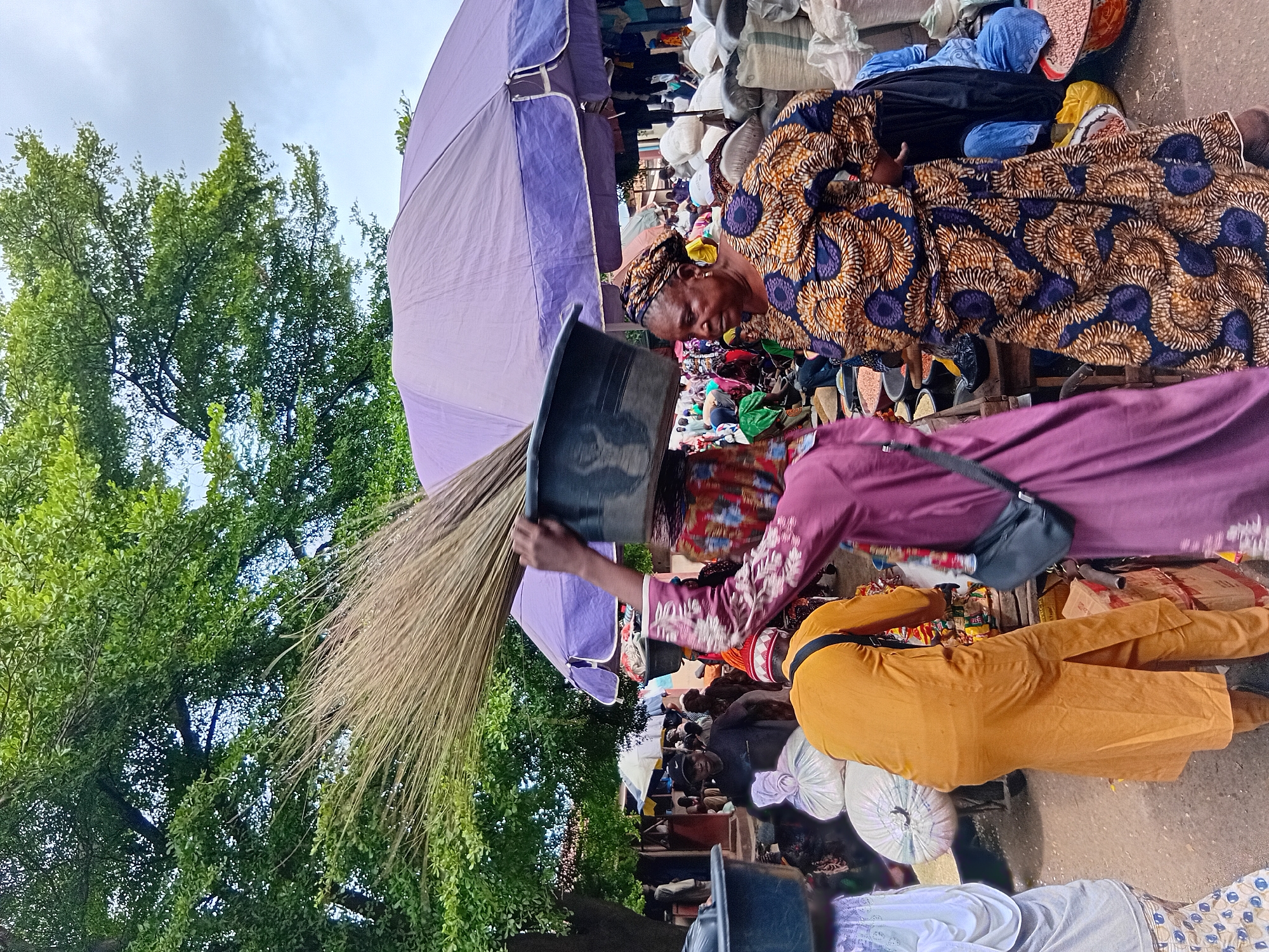 The image depicts a bustling outdoor market scene in Lokoja, Nigeria, under a cloudy sky and shaded by large trees and umbrellas. In the foreground, a woman carrying a large basin filled with what appears to be broom material balanced on her head walks from left to right. She is wearing a vibrant, patterned dress with blue and orange motifs and a headscarf. To her right, another woman, also wearing a colorful, patterned dress and headscarf, is looking towards the left.
Behind these women, the market is filled with people and goods. A man in an orange tunic and pants stands to the left, facing away from the camera. Numerous stalls are visible, displaying sacks of grain and other commodities. Large purple and white umbrellas provide shade. People are seen interacting, browsing, and carrying items. The ground is a dusty, reddish-brown earth. The overall impression is one of lively commerce within a traditional market setting.