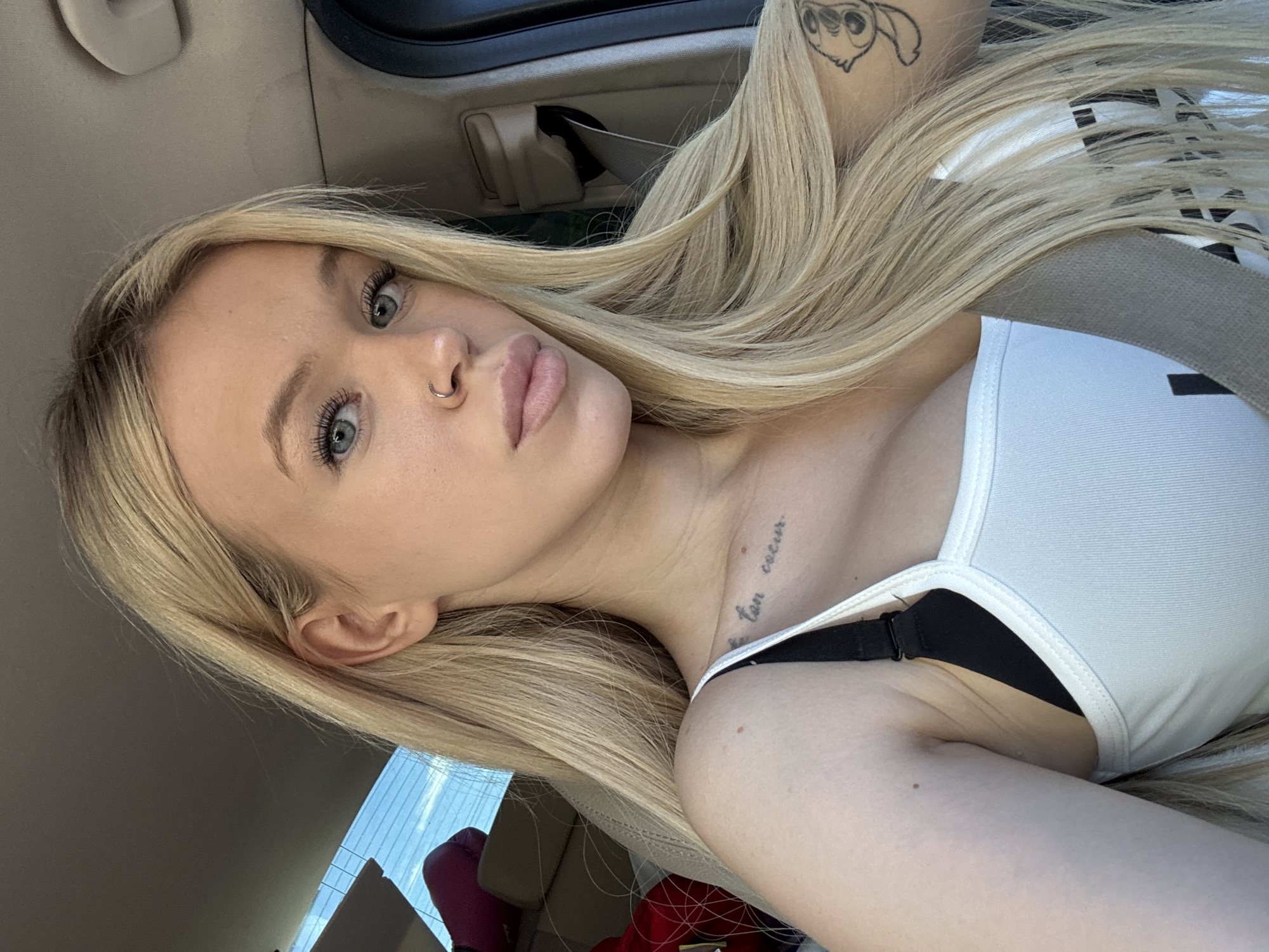 The image is a close-up, vertically oriented selfie taken from the perspective of someone in the passenger seat of a car. The primary subject is a young woman with long, blonde hair and light blue eyes. She is wearing a white tank top and a black bra. Her head is tilted, and her gaze is directed towards the camera. A small nose ring is visible, and a tattoo of a cartoon character, possibly Stitch from Disney's Lilo & Stitch, is on her upper arm. Another tattoo, a script of text, is visible on her collarbone.
The background suggests the interior of a car, with a seatbelt buckle and part of the car's interior trim visible. Through the car window, a blurry background with what appears to be a bright sky and some red objects can be seen. The lighting suggests it is daytime, likely sunny, casting a soft glow on the subject's skin. The overall mood conveyed is casual and personal. The provided location context is Saint Petersburg, Russia, but there are no specific visual cues within the image to confirm this.