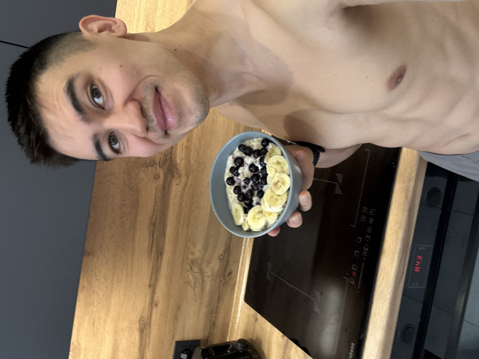 A shirtless man holds a bowl of oatmeal with blueberries and banana slices. He stands in a kitchen with wood counters and a black cooktop. The cooktop's display reads "8:43." The man smiles at the camera while holding his breakfast. The background features a gray wall.