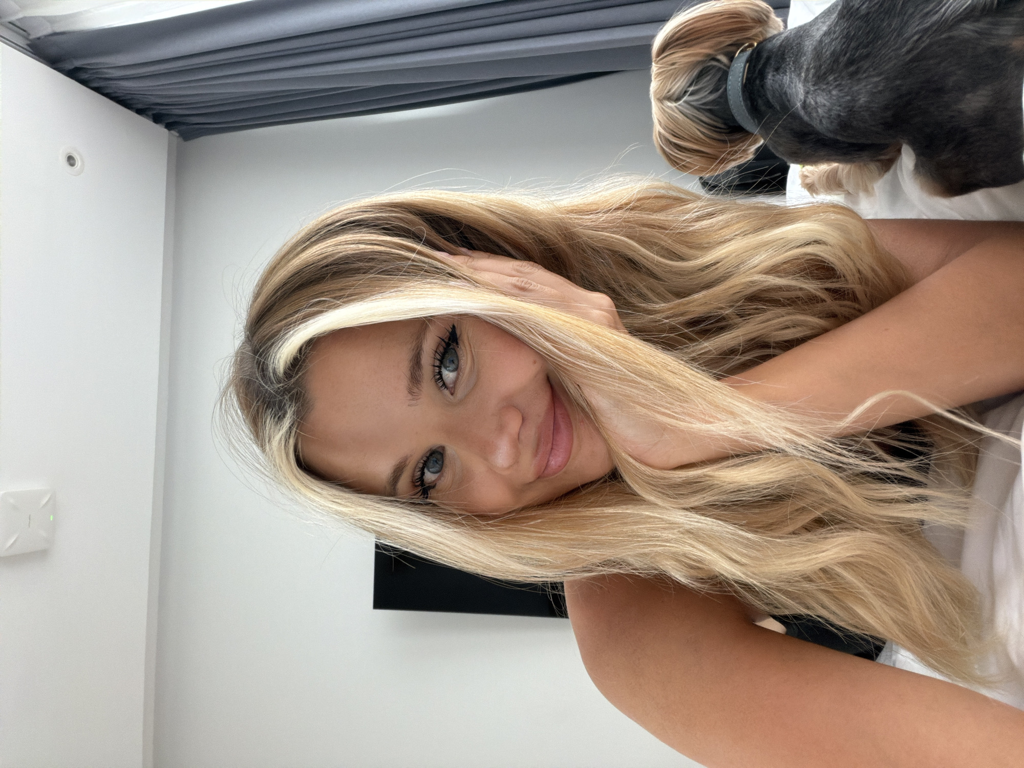 A young woman with long, blonde, wavy hair is lying down, with her head tilted. She is looking at the camera with a gentle smile. Her eyes are blue and she has light makeup on. Her left hand is touching her hair, near her face. A small dog, mostly black with some grey, is resting its head on her back, near her shoulder. The background appears to be a bedroom, with white walls and a grey curtain or headboard above. The lighting suggests it could be daytime. There are no visible text elements in the image.