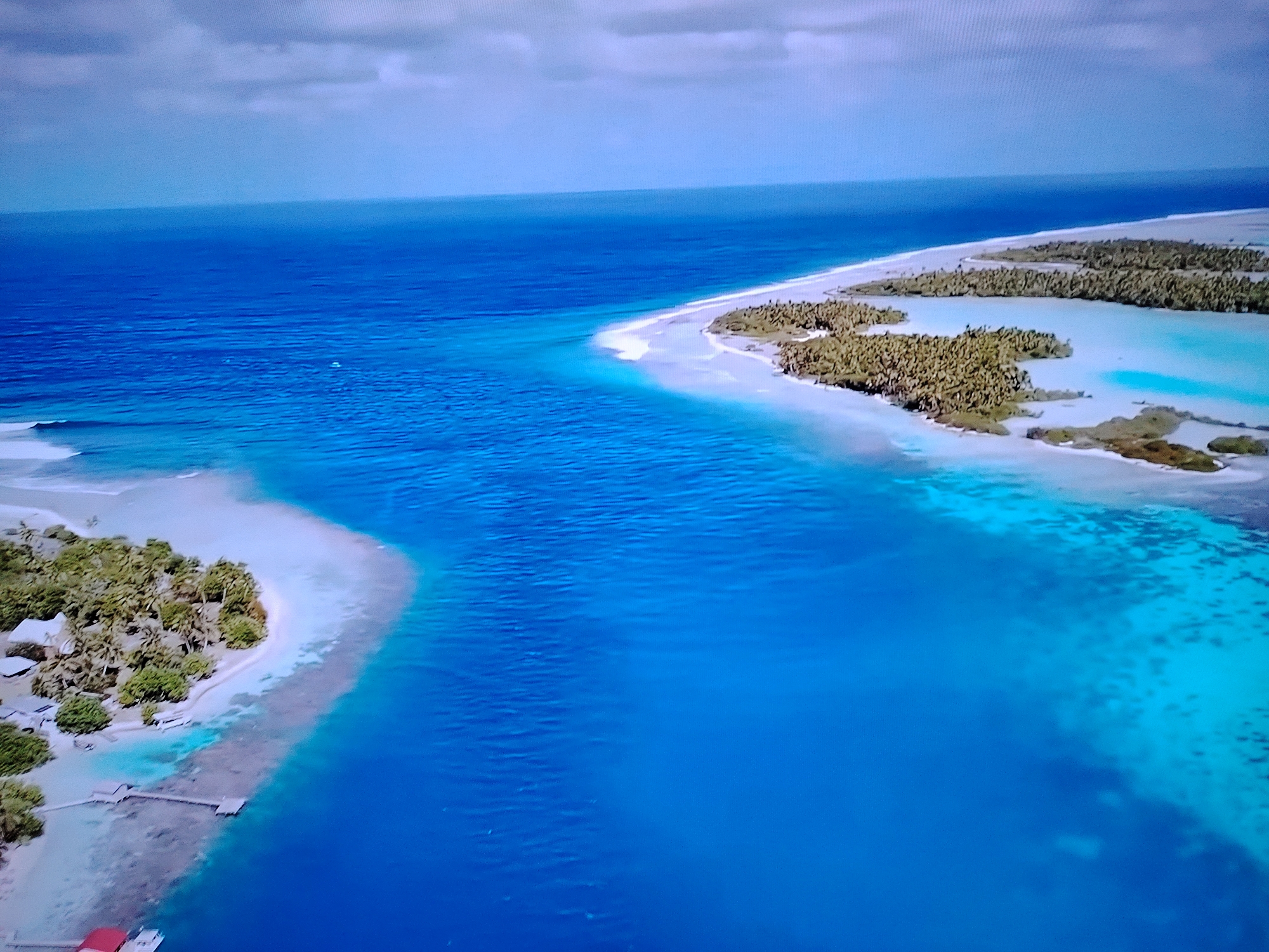 This is an aerial view of a tropical atoll with clear turquoise waters and lush green vegetation. Several small islands, covered in palm trees, are scattered throughout the lagoon. The islands have white sandy beaches and the water around them is shallow and a vibrant blue, indicating coral reefs beneath the surface. In the foreground, a small pier extends into the water from one of the islands, with a building and some vegetation visible on the shore. A boat with a red roof is docked at the end of the pier. The sky above is partly cloudy with a soft, hazy light. There are no visible people in the image. This scene is not located in Huntingdonshire, United Kingdom, as it depicts a tropical environment.