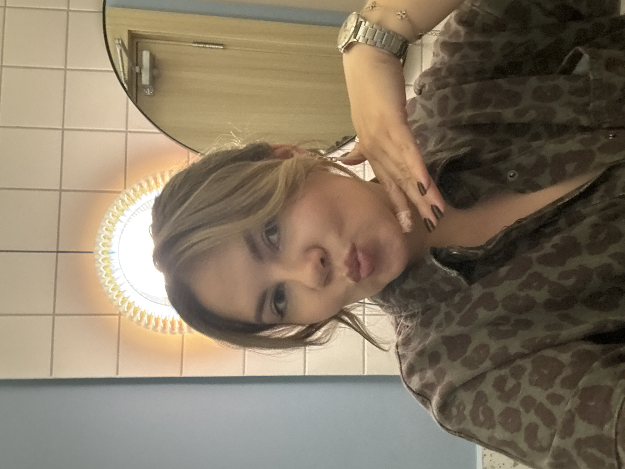 The media file shows a portrait of a young woman in a bathroom. She is leaning towards the camera, with her right hand touching her cheek. She has blonde hair with bangs, and is wearing a leopard-print jacket. A silver watch is visible on her left wrist. In the background, there are white tiled walls, a circular mirror with a bright light behind it, and a door. The overall lighting suggests it is daytime. The scene appears to be a private bathroom, possibly in a home or public restroom. The woman's expression is a pout, suggesting a playful or flirtatious mood. No visible text is present in the image.