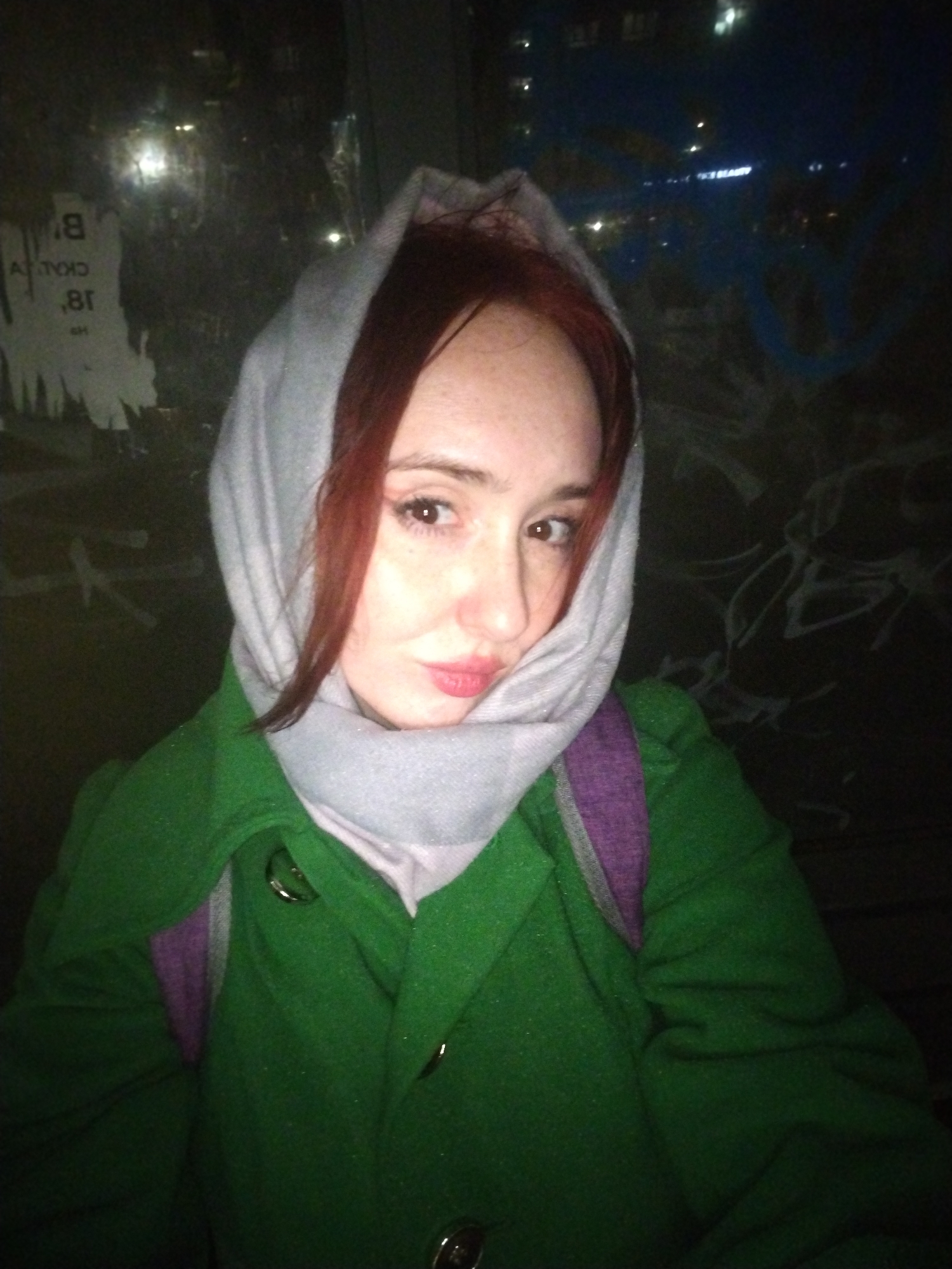 A young woman is posing for a selfie at night in Izhevsk, Russia. She is wearing a green coat, a light gray headscarf, and a purple backpack. The background includes graffiti and posters on a wall, along with blurred city lights in the distance. A sign with partial letters "CKAL" is visible on one of the posters. The woman has reddish-brown hair and is looking directly at the camera with a slight pout.