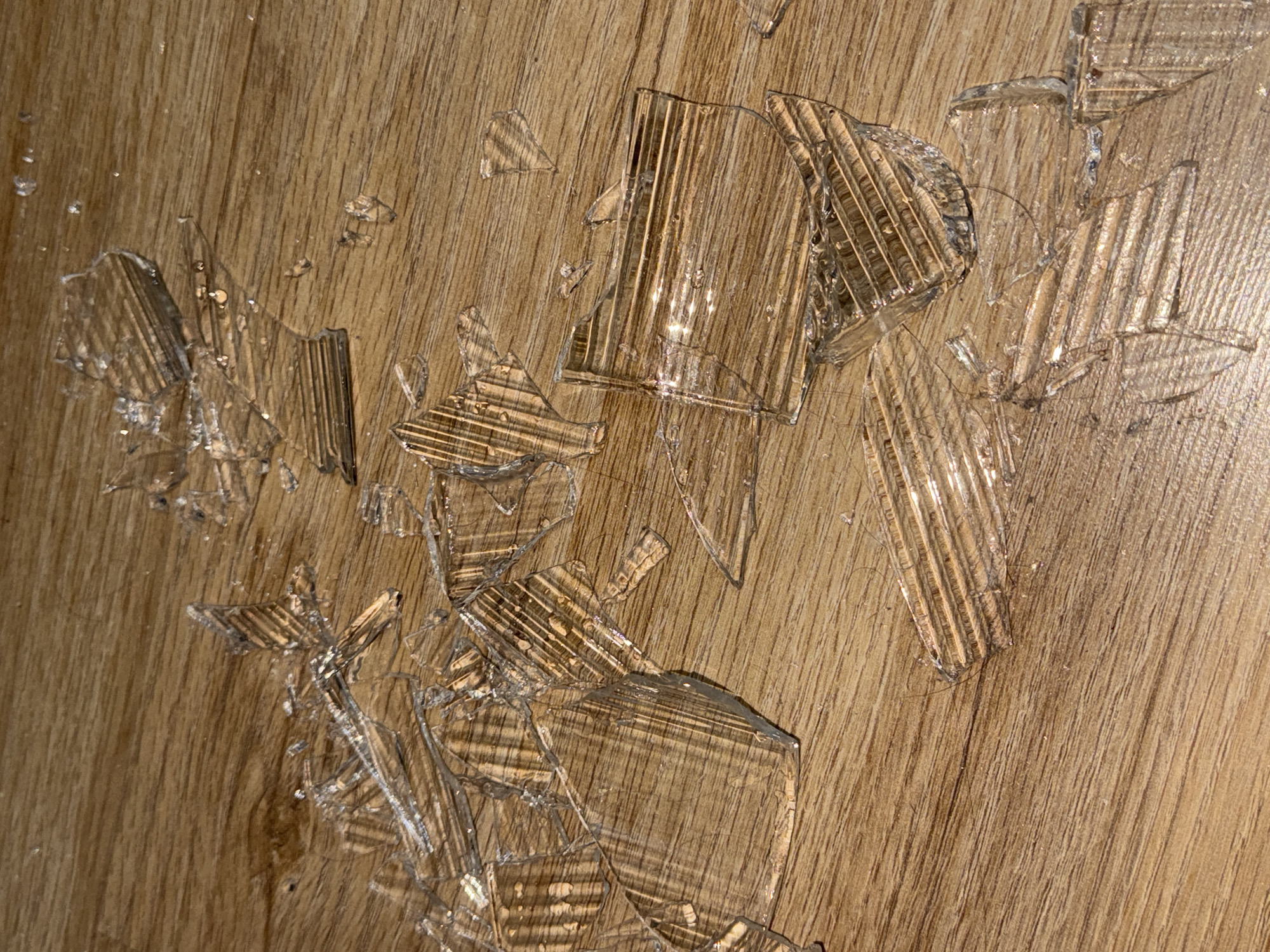 The scene displays numerous clear glass fragments scattered across a light brown, wood-patterned floor in an indoor environment, consistent with a location in Perm, Russia. The glass shards vary in size and irregular shape, with many pieces exhibiting a distinct ribbed or corrugated texture on one surface. Reflections of an unseen light source are visible on the fractured glass. The floor appears to be wood laminate or hardwood, exhibiting a horizontal grain. No individuals, additional objects, or active events are depicted.
