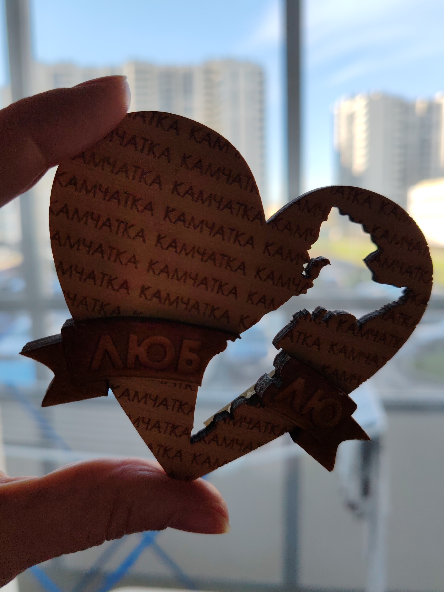 A person's hand holds a heart-shaped wooden souvenir against a bright, out-of-focus outdoor background. The souvenir is designed as a heart split into two pieces, currently held slightly apart. The larger left piece and smaller jagged right piece are patterned with repeating Cyrillic text: "КАМЧАТКА" (KAMCHATKA), referring to the Kamchatka Peninsula in Russia.
The wooden pieces show darkened edges, indicative of laser cutting. A small, bird-shaped cutout is visible near the break point on the left piece. At the bottom of each piece, a banner-like section contains embossed Cyrillic letters. The left banner reads "ЛЮБ" (LYUB), and the right reads "ЛЮ" (LYU). When combined, these form "ЛЮБЛЮ" (LYUBLYU), meaning "I love" in Russian, conveying the sentiment "I love Kamchatka."
The background, seen through a window, features blurred multi-story residential buildings against a clear, blue sky, indicating bright daylight. A window frame and a vertical mullion are discernible. The bright, clear weather suggests the photo was taken on a sunny day, likely in Saint Petersburg, Russia. The object is a personalized memento or keepsake.