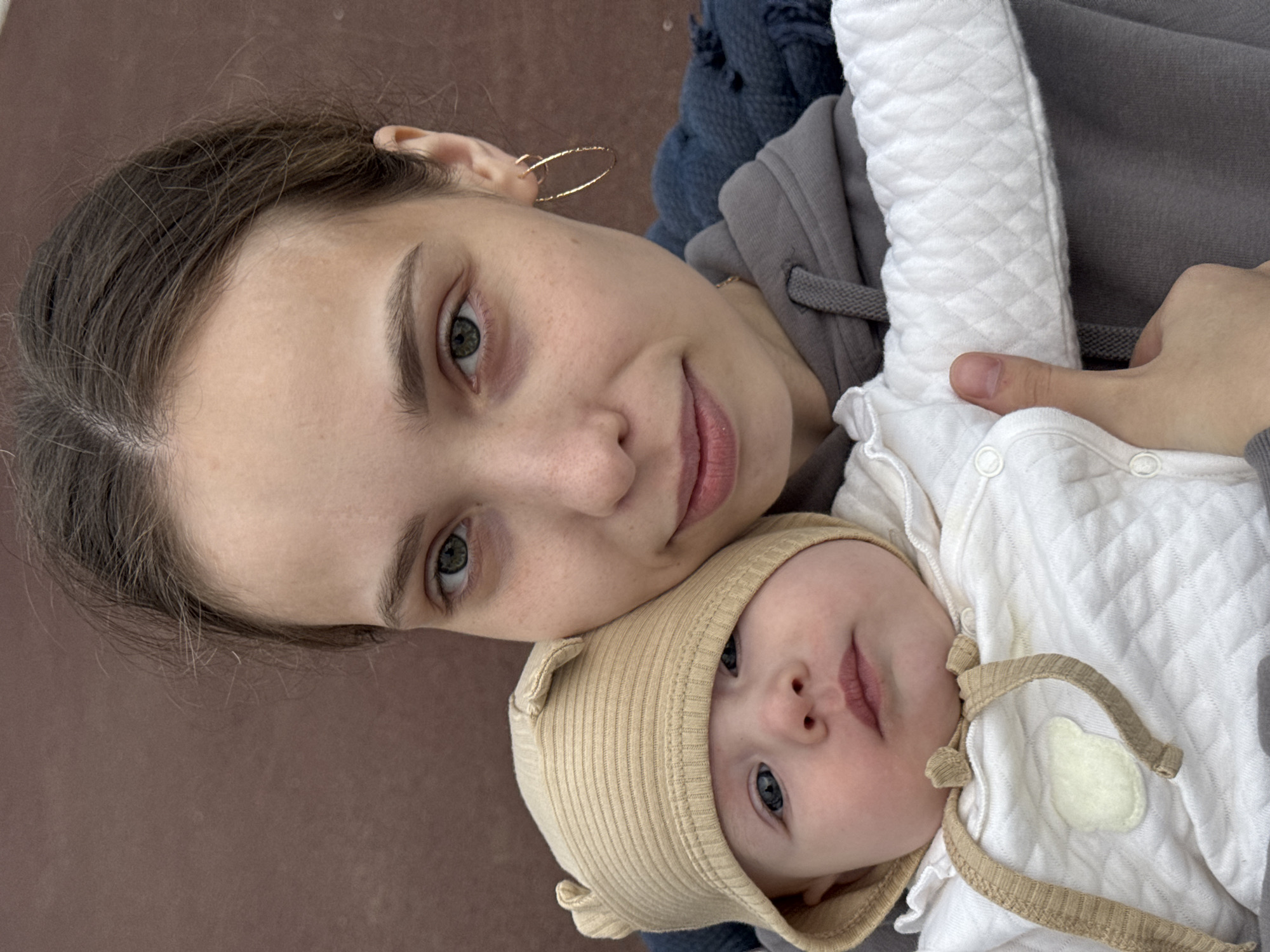 An overhead, close-up selfie shows a young woman holding a baby. The woman has light skin, dark brown hair pulled back, and green eyes. She is wearing a grey hooded sweatshirt and a dark blue scarf. The baby, facing the camera, is wearing a light brown ribbed hat with ear details and a white quilted onesie with a white applique patch in the shape of a cloud. The baby has blue eyes and light skin. The background is a solid, dark brown surface. The image appears to be taken outdoors.