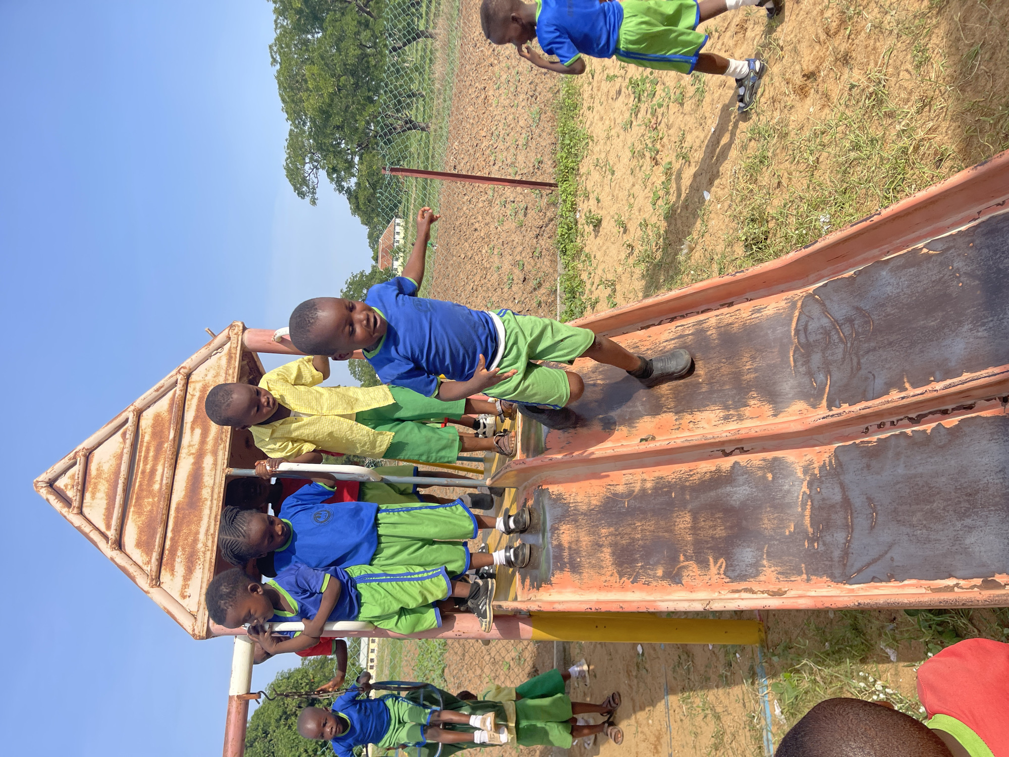 Akwanga, Nigeria. Several children in school uniforms (blue shirts with green shorts) play on and around a weathered metal slide and a swing set on a sunny day. The playground is situated on sandy soil with sparse grass. A few children are in the structure at the top of the slide. Another is exiting the slide. One child stands a short distance away from the slide, near a fence and an agricultural field. One child sits on the swing.