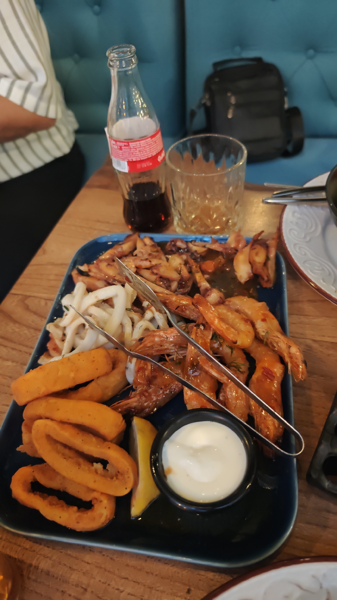 A tray of seafood is placed on a wooden table. The tray contains fried calamari rings, white calamari strips, fried octopus pieces, and prawns. A lemon wedge and a small bowl of white sauce are also on the tray. A pair of metal tongs is placed on top of the food. A glass of amber liquid and a small glass Coca-Cola bottle are also visible. A person wearing a striped shirt is partially visible on the left side of the frame. A black bag rests against a blue upholstered wall in the background. This could be in a restaurant or a similar establishment in Burgas, Bulgaria.