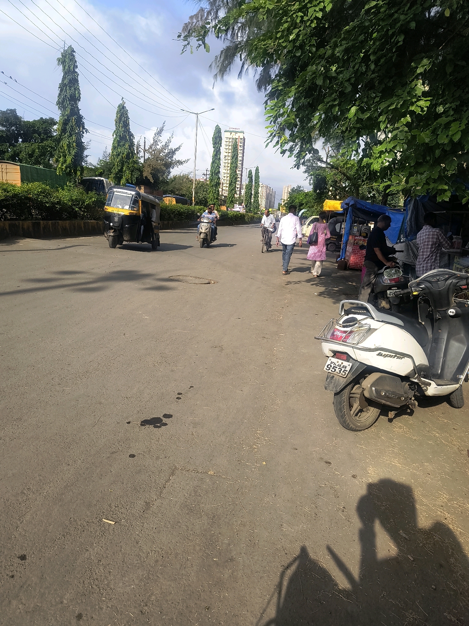 The image captures a street scene in Vasai-Virar, India, with a focus on a road that appears to be a thoroughfare. In the foreground, a white scooter is parked, its license plate reading "MH.48.DN.9535". Further down the road, an auto-rickshaw is in motion, its yellow and black color scheme prominent. A person is riding a scooter behind the auto-rickshaw. On the sidewalk, several individuals are visible, some near makeshift stalls or shelters covered with tarps. One person is seen cycling, and another is walking. The environment is characterized by buildings in the background, including what appears to be a high-rise structure. Lush green trees line one side of the street, casting shadows on the road. The sky is partly cloudy, with a bright, daylight illumination. The road surface shows signs of wear and tear, with some dark oil stains scattered across it.