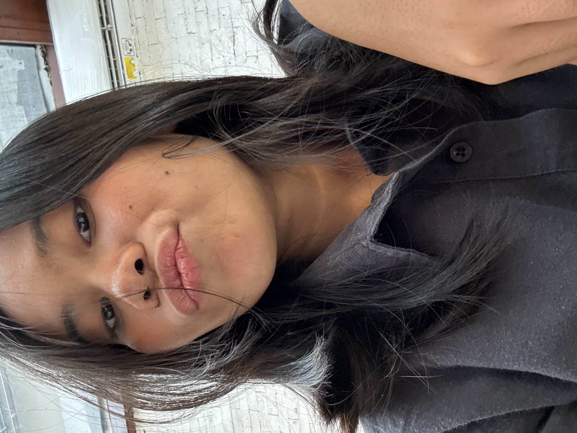 In a close-up, head-and-shoulders indoor shot, a young woman with long, dark hair pulled back slightly from her face puckers her lips. She is wearing a dark grey collared shirt, the buttons visible. Her skin appears smooth with a natural tone, and a small mole is visible on her left cheek. The background consists of a white brick wall and what appears to be an air conditioning unit. The lighting is soft and diffused. The location is identified as South Jakarta, Indonesia.