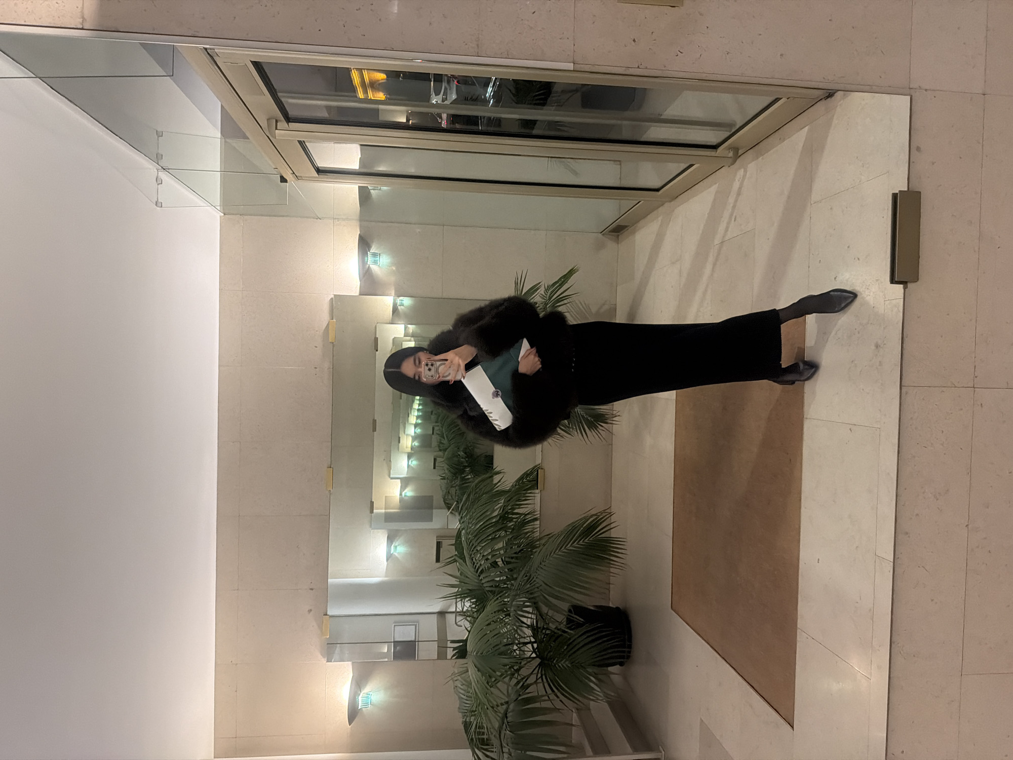 A photograph, taken in a well-lit interior space with neutral-toned walls and tiled flooring, shows a person reflected in a large mirror. The space appears to be a lobby or entrance area. The person, seemingly female, is dressed in black attire, including a long skirt or dress and what appears to be a fur coat or jacket. She is holding a mobile phone in her right hand, seemingly taking a selfie, and a green and white object in her left hand. In front of the mirror is a large potted palm tree. The mirror reflects a door or window frame above the person and repeating hallway with sconces and what may be a seating area. The light fixtures on the walls are partially visible in the reflections. The mirror has a frame that blends with the neutral colors of the hallway. Based on the architectural style and overall ambiance, this location appears to be in Paris, France.