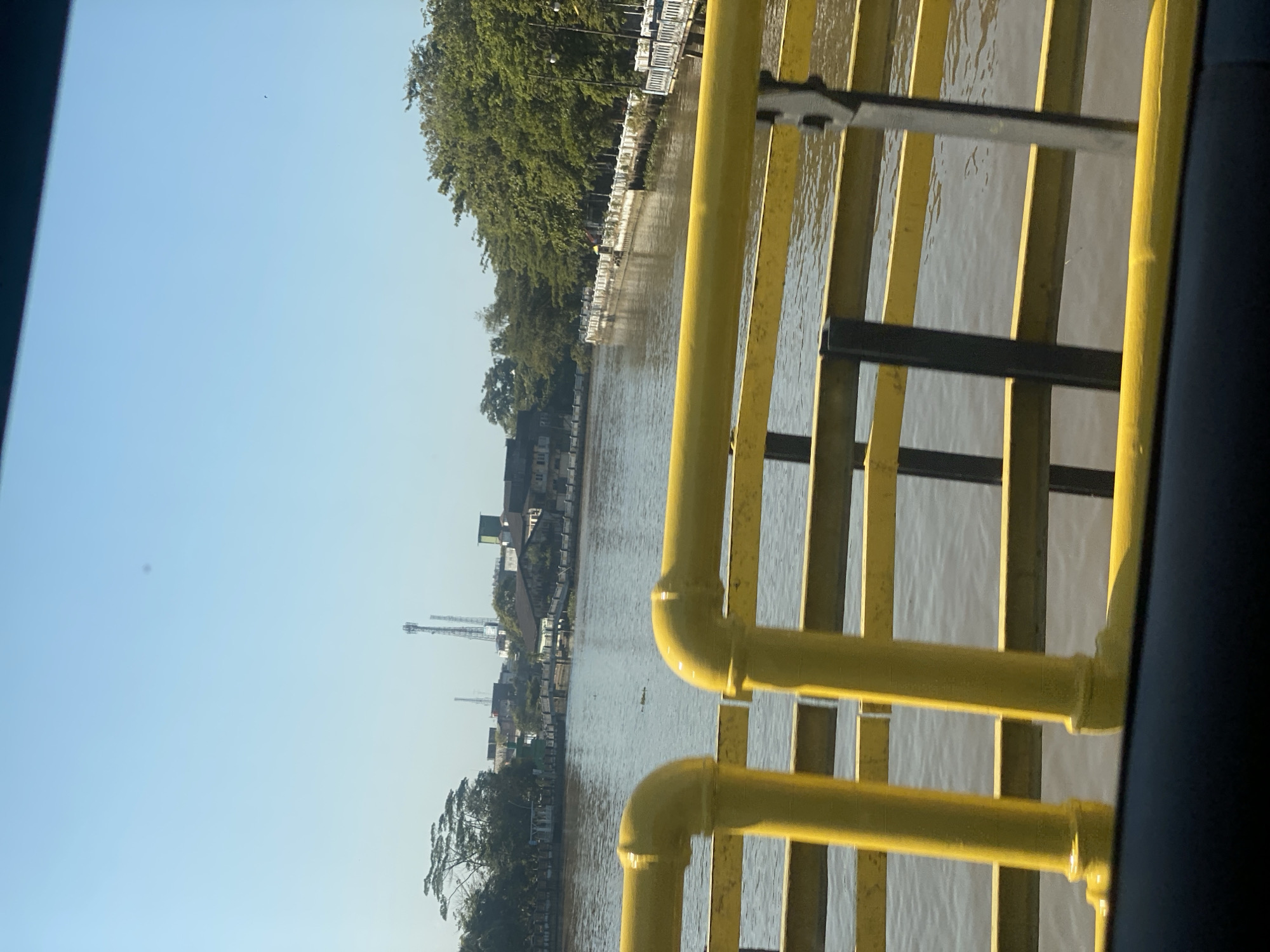 This is a photograph taken from a bridge overlooking a river in Banjarmasin, Indonesia. The foreground is dominated by a yellow railing, likely part of the bridge structure, with thick vertical pipes and horizontal bars. The river itself occupies the mid-ground, its surface rippling gently. On the far bank of the river, a tree-lined promenade is visible, featuring a white railing with ornate posts and lampposts. Buildings and structures, including what appears to be a mosque with a green dome, are nestled among the trees and further in the distance, two prominent cellular towers rise into the clear blue sky. The overall impression is of a sunny day in an urban riverside setting.
