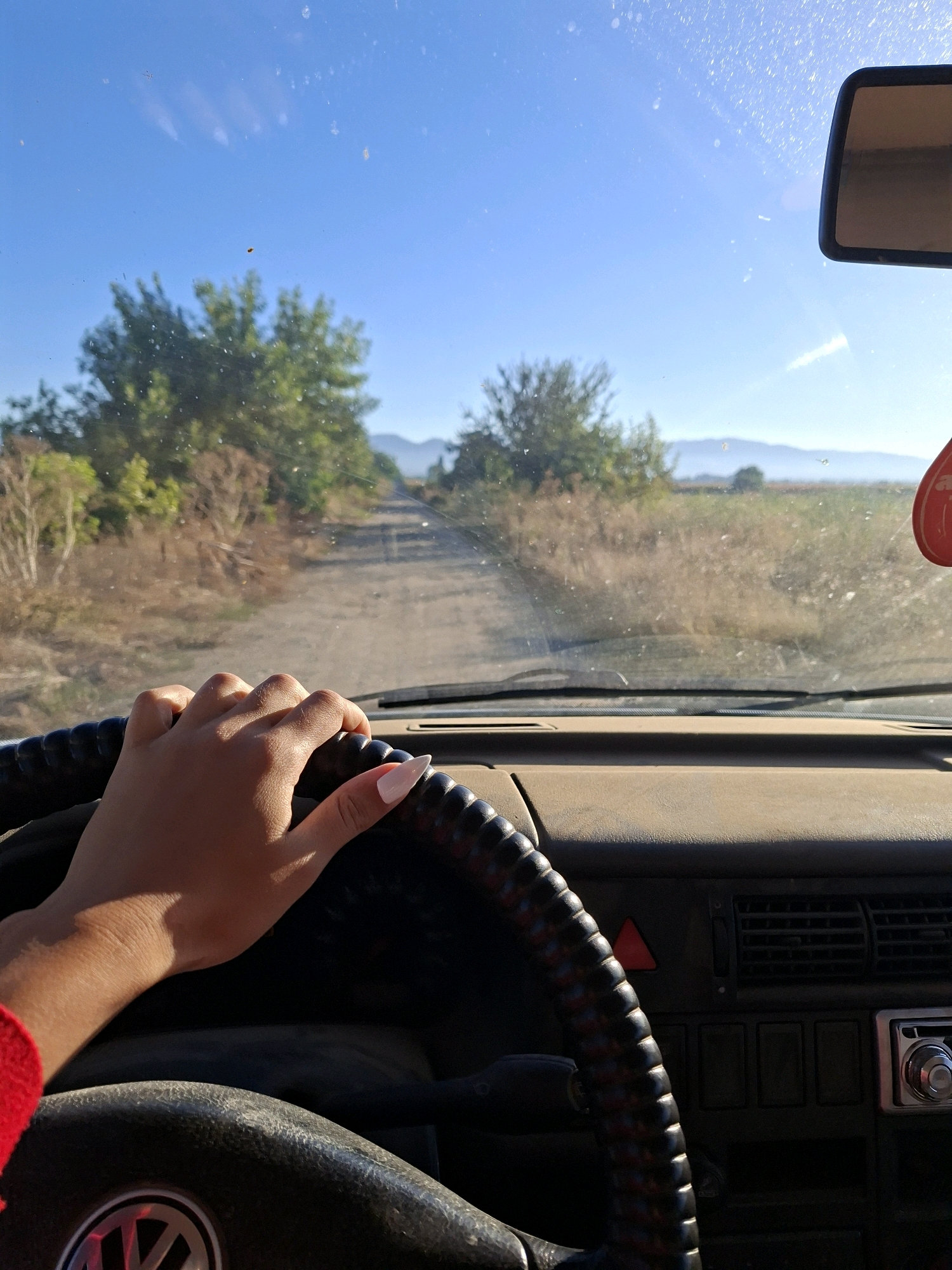 A first-person perspective shot shows a hand on a steering wheel inside a vehicle. The hand is resting on the top of the steering wheel, with the palm facing down and fingers curved. The fingernails are painted with a light color. The dashboard is visible, with a car radio and air vents. Outside the vehicle, a dirt road stretches into the distance, flanked by dry grass and trees. Mountains are visible in the background under a clear blue sky. A red car air freshener hangs from the rearview mirror.