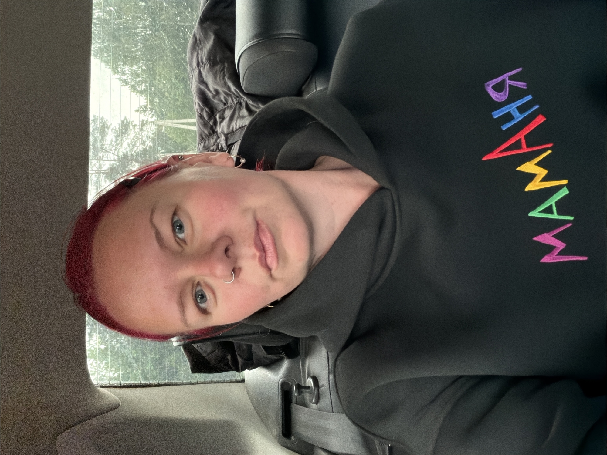 A woman with reddish-pink hair, blue eyes, and a silver hoop nose piercing is featured in a portrait-oriented photograph taken inside a car in Chaikovsky, Russia. The image is rotated 90 degrees clockwise. She looks directly at the camera with a neutral expression, showing a slight flush on her cheeks. She is wearing a black hooded sweatshirt with the word "MAMAMA" embroidered in colorful, stylized letters across the chest. The letters are purple, blue, red, yellow, green, and pink.
The interior of the car is visible around her, including a light-colored headrest to her left, a darker seat or headrest behind her to the right, and a seatbelt buckle near the bottom. Through the rear window, an outdoor scene with numerous trees and green foliage is visible. The lighting is diffused, suggesting an overcast or rainy daytime. A dark, crumpled jacket or piece of clothing is also visible on the seat behind her.