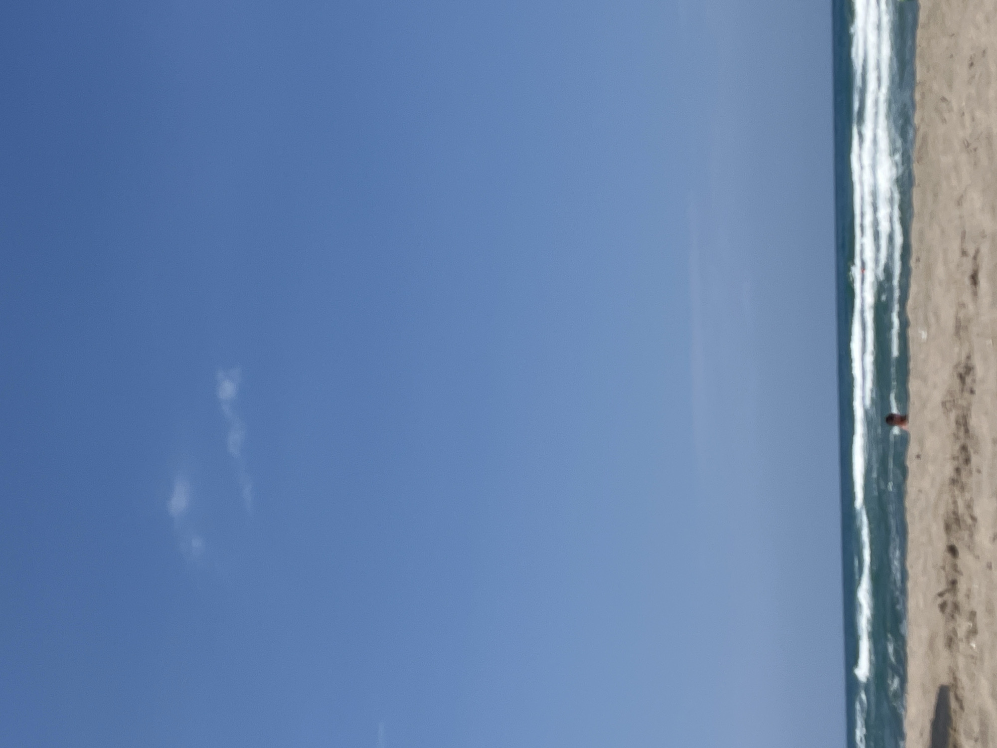 The image depicts a beach scene in Ahtopol, Bulgaria. The sky is a clear, bright blue with a few wispy clouds. The sea is a deep blue-green with white-capped waves rolling towards the shore. The beach is sandy and appears to be mostly empty, with some slight undulation. In the middle ground, partially submerged in the water, a person's head is visible. The person appears to be swimming or wading. In the foreground, the sand is closer to the viewer, with some areas of shadow suggesting either dunes or footprints. The overall impression is one of a peaceful, sunny day at the beach.