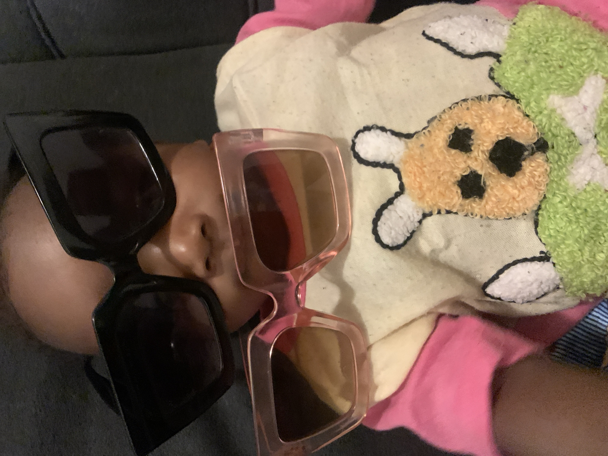 A child is resting on what appears to be a gray couch, positioned next to a light-colored blanket featuring a sheep or lamb design. The blanket is a pale cream color with patches of fluffy, light-green material. The child is wearing a pink garment. The child's face is partially obscured by two pairs of sunglasses. One pair is black and angular, while the other is pink and rectangular.
There is no information in the image to indicate this photo was taken in Barakin Akawo, Nigeria.