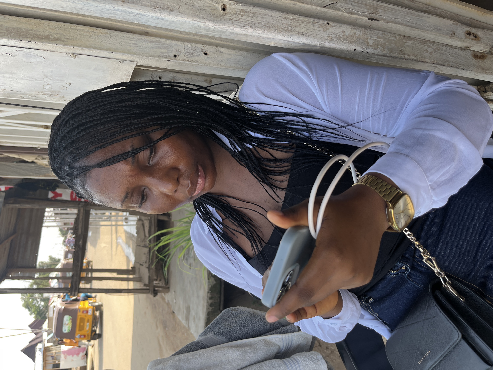 A medium shot, taken outdoors, shows a young Black woman with braided hair sitting and looking down at her phone. She is wearing a white open button-down shirt over a black tank top and blue jeans. A gold watch is on her left wrist, and a black quilted purse with a gold chain is on her lap. The woman is holding a light gray phone in her hands. The background shows a street scene with a yellow auto-rickshaw in the distance and some weathered wooden structures. The image appears to be taken in a market or roadside area in Abuja, Nigeria.