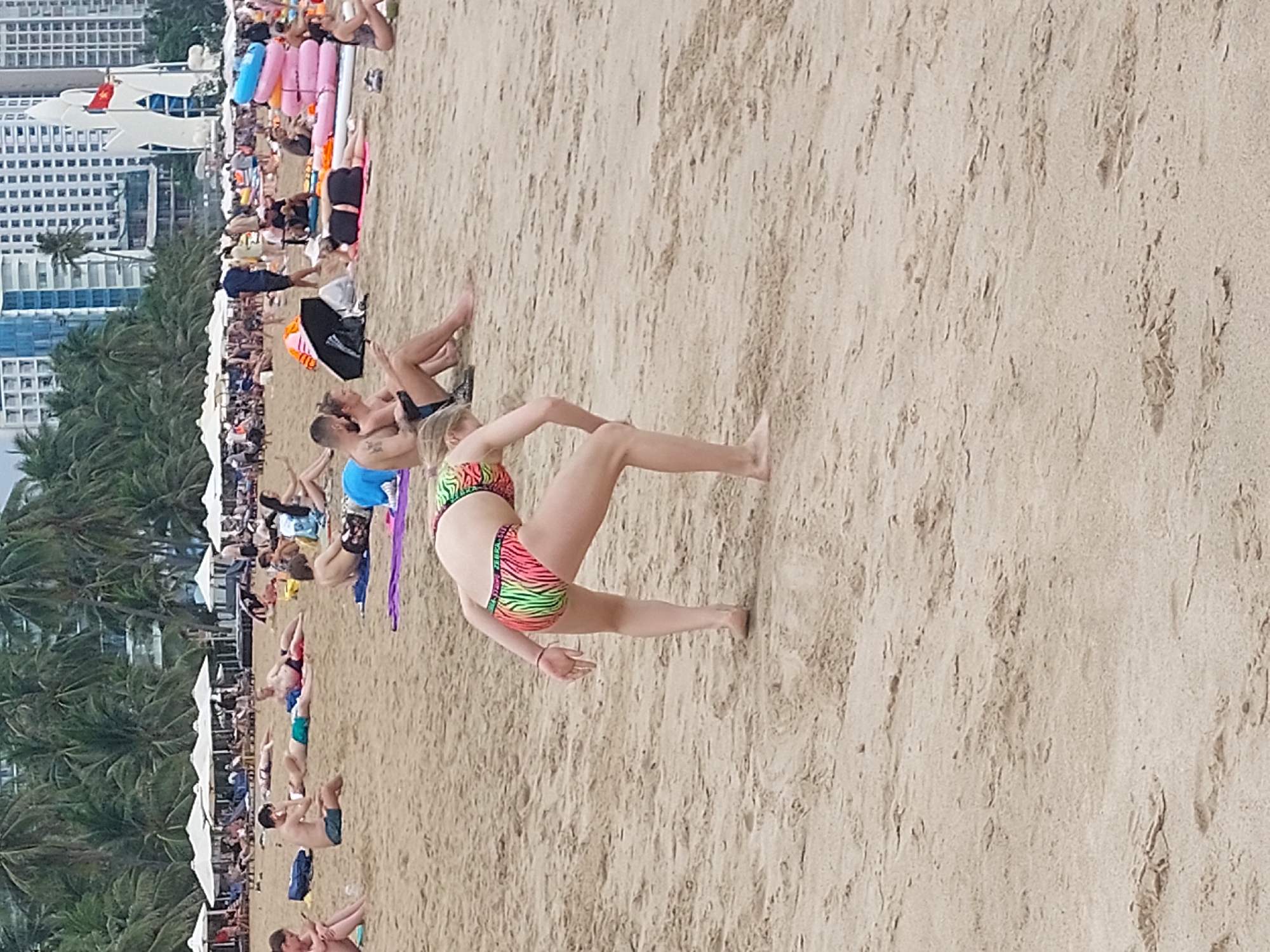This media file depicts a busy beach scene in Nha Trang, Vietnam. The foreground features a woman in a vibrant, multi-colored bikini striking a yoga pose on the sand. Her body is positioned in a dynamic stance, with one leg extended and arms outstretched, showcasing her flexibility. The sand is the dominant element in the mid-ground and background, with visible textures and patterns created by the natural environment and human footprints.
Further back on the beach, numerous people are engaged in various leisure activities. Some are lying down on towels or in the sand, possibly sunbathing or relaxing. Others are sitting or standing, interacting with each other. In the distance, a row of white beach umbrellas provides shade, suggesting a organized area for beachgoers. Beyond the umbrellas, a line of lush palm trees forms a natural boundary, with the silhouettes of tall buildings visible behind them. The architecture of these buildings, appearing modern and high-rise, indicates an urban setting adjacent to the beach.
The scene is bathed in what appears to be natural daylight, likely during the daytime given the presence of sunbathers and the overall brightness. The weather seems to be fair and conducive to beach activities. The presence of numerous people suggests a popular tourist destination.
There is no visible text within the media file that provides any specific information about the event or location. The overall atmosphere appears relaxed and casual, consistent with a beach day. The image captures a slice of life at a popular Vietnamese beach, highlighting human interaction with the natural environment and recreational pursuits.