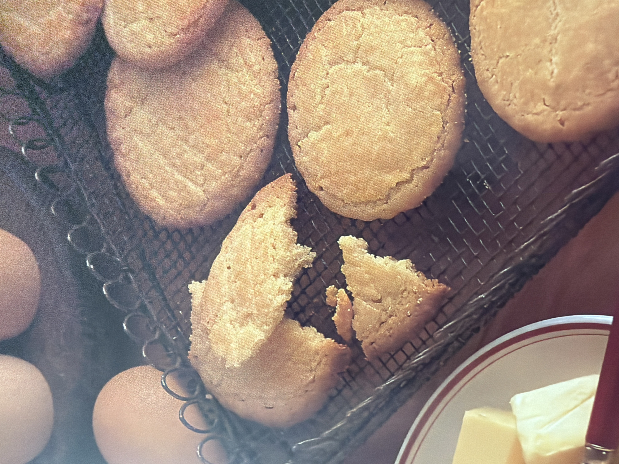 The image displays a close-up view of several golden-brown cookies resting on a metal cooling rack. Some of the cookies are whole, while one in the foreground is broken into two pieces, revealing a crumbly interior. The cookies have a slightly uneven surface, characteristic of homemade baked goods.
In the background and to the left of the cookies, several brown eggs are visible. Partially in the lower right corner of the frame, a plate with a red and white rim holds two blocks of pale yellow butter, and a red knife handle protrudes from the right side of the plate.
The lighting suggests an indoor setting, possibly a kitchen. The overall scene evokes a sense of rustic baking and the preparation of ingredients for a meal or dessert. There is no visible text in the image. The image is a still photograph, and no specific time of day, weather conditions, or explicit location cues are discernible. The focus is on the textures and colors of the food items.
