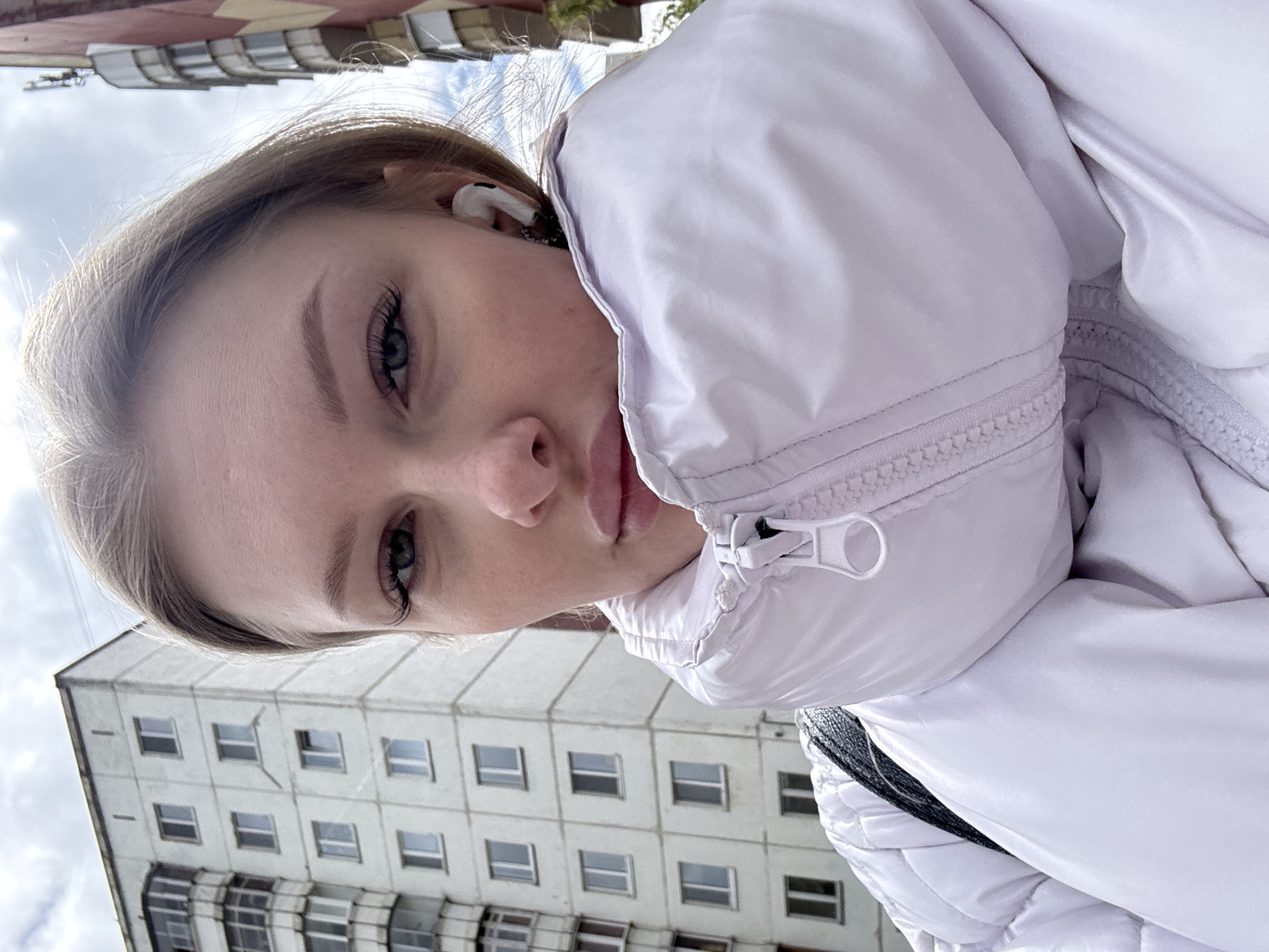 A young fair-skinned woman with light brown hair and gray-blue eyes is wearing a white puffer jacket and an earbud. She is in an urban environment, with a tall, light-colored apartment building behind her. The sky is overcast. The location is likely in Perm, Russia, based on the style of architecture of the building and the general weather patterns.
