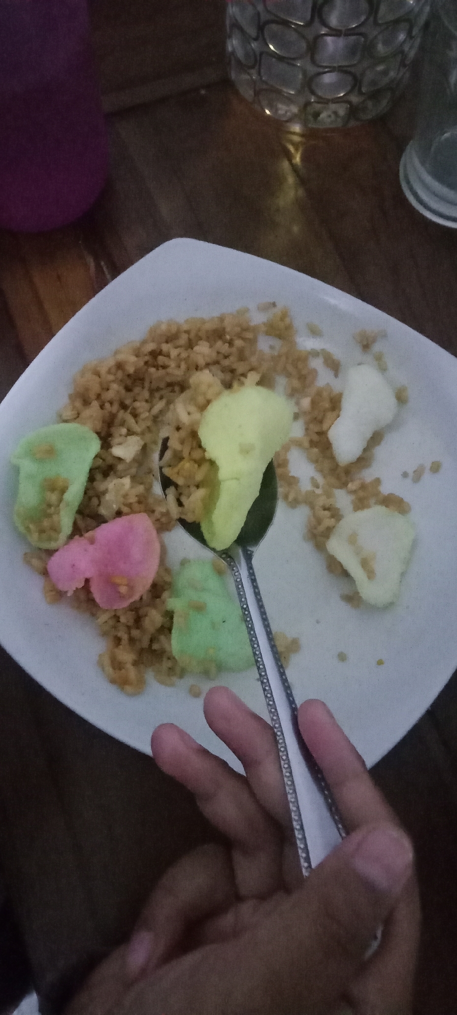 A close-up, eye-level, full shot captures a plate of fried rice with colorful crackers and a hand holding a spoon. The fried rice appears to be a light brown color, with visible grains and possibly some small pieces of egg or vegetables mixed in. Scattered on top of the rice are several puffy, brightly colored crackers. There are two light green crackers, one pink cracker, one light yellow cracker, and two white crackers. A silver spoon with a decorative rim is placed in the center of the plate, partially covered by the rice and crackers. A human hand, with visible fingernails and knuckles, is seen in the foreground, holding the handle of the spoon. The hand appears to be that of an adult. The plate is white and oval-shaped. The background consists of a dark wooden surface, likely a table. To the left of the plate, a portion of a purple cylindrical object is visible, possibly a cup or bottle. In the upper right corner, a blurred, patterned object, likely a decorative glass or container, is partially visible. The lighting is dim, creating shadows and highlighting the texture of the food and the spoon. The scene is likely indoors, possibly in a home or restaurant setting. The image is taken in Krinjing, Indonesia.