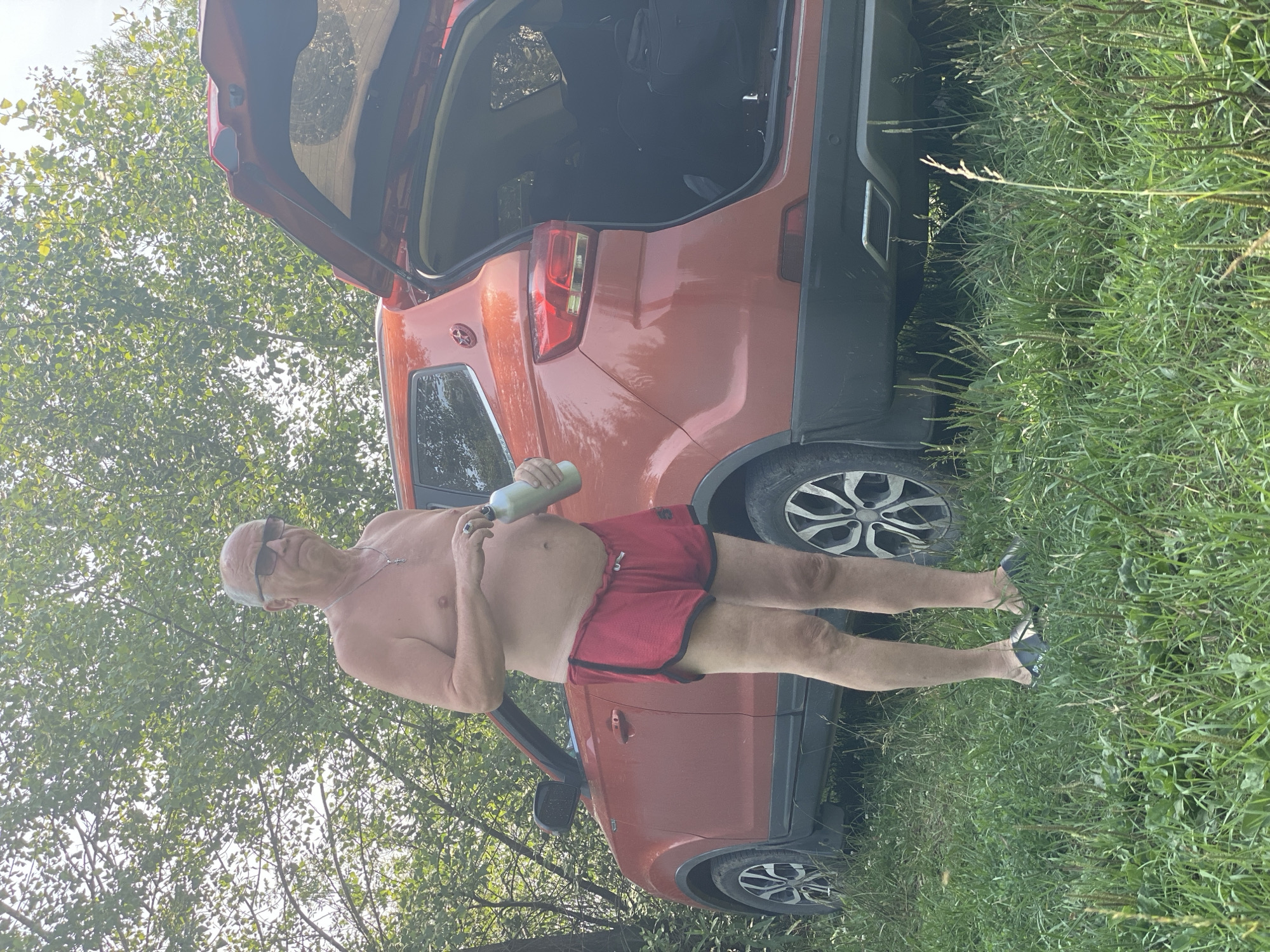 An elderly Caucasian man, shirtless and wearing red athletic shorts and black flip-flops, stands next to a red SUV with its rear hatch open. He is holding a silver water bottle. The scene appears to be outdoors, in a grassy area with trees in the background. The car is parked on what appears to be a dirt or gravel surface, suggesting a rural or undeveloped area rather than a typical city street. There is no visual information in the image that would indicate the specific location as Novokuznetsk, Russia.